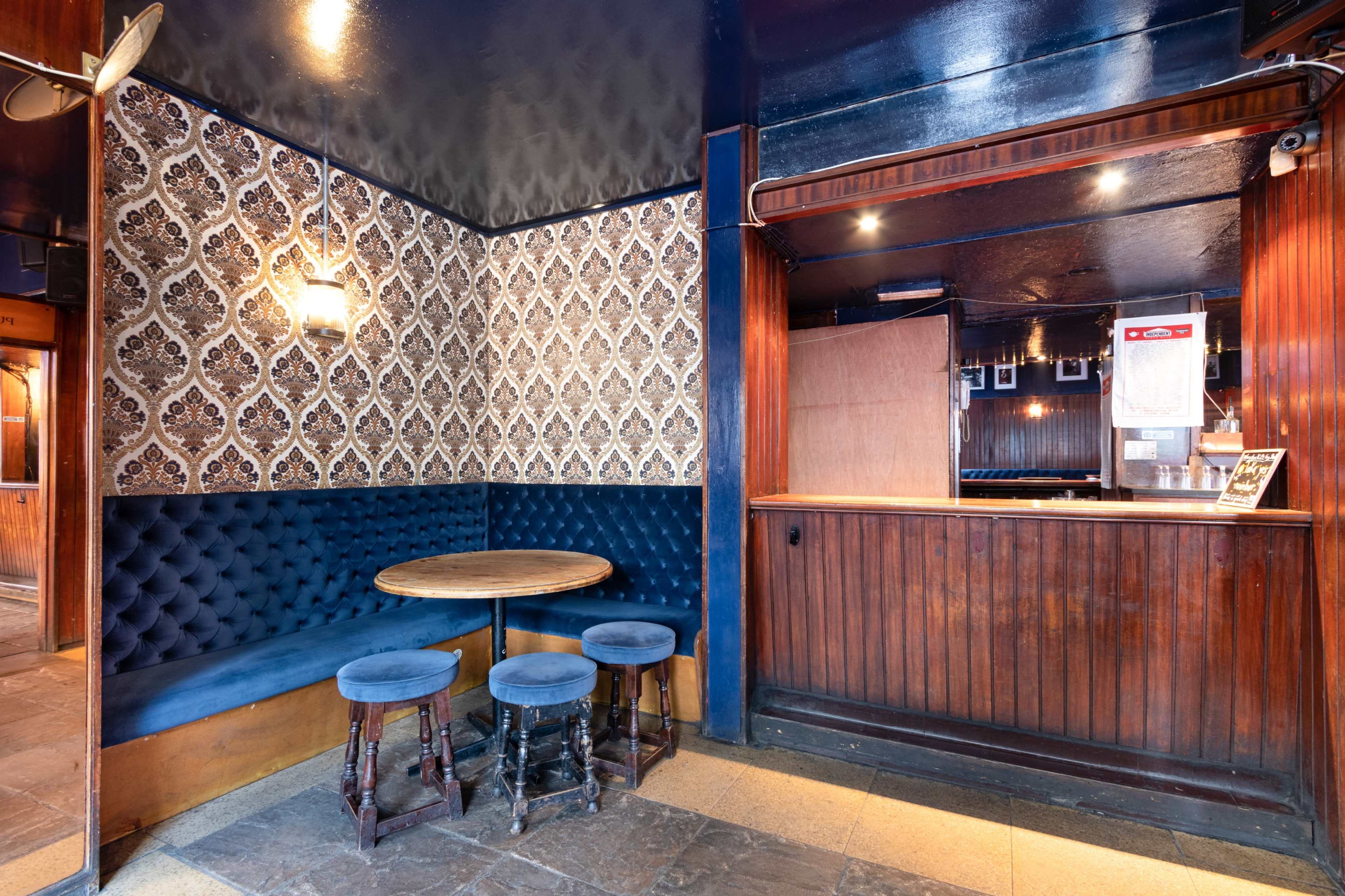 The image shows a corner of a dimly lit bar featuring a blue velvet booth, a round wooden table, and a wooden bar counter with decorative wallpaper in the background.