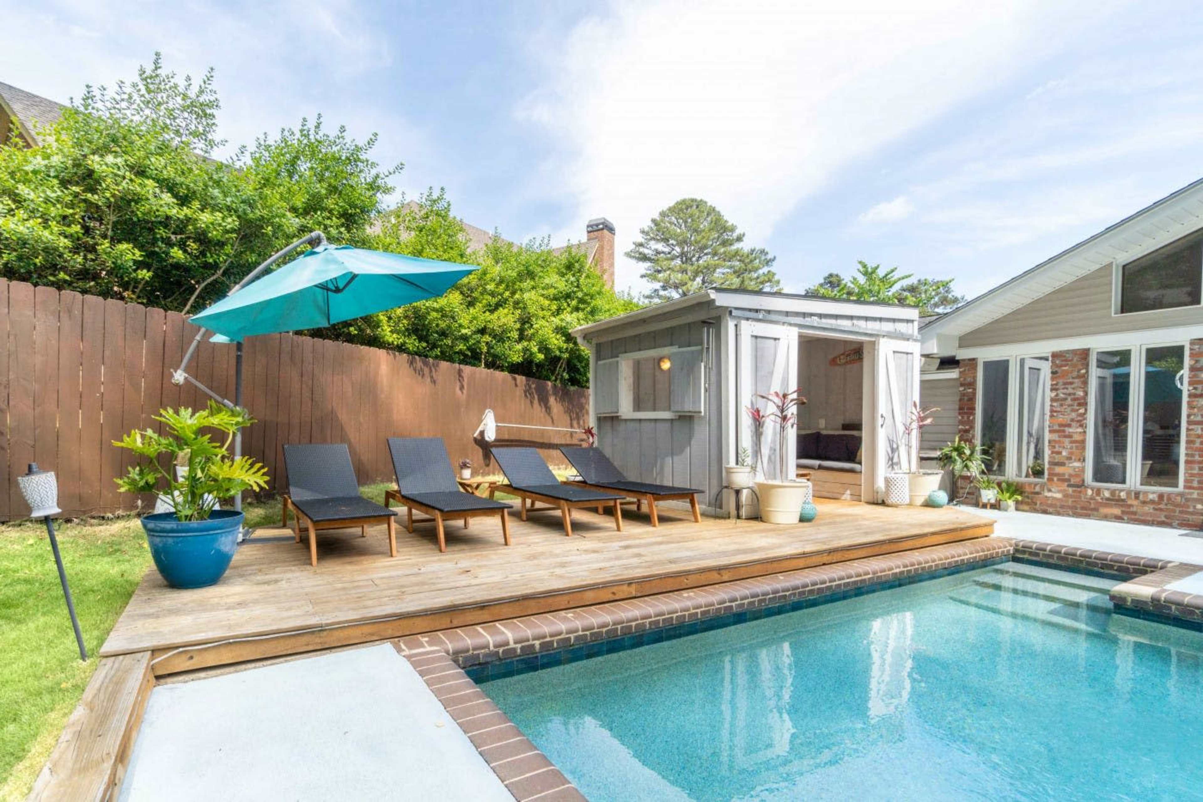 A wooden deck with three lounge chairs, an umbrella, and a small storage shed beside a swimming pool surrounded by greenery and a wooden fence.