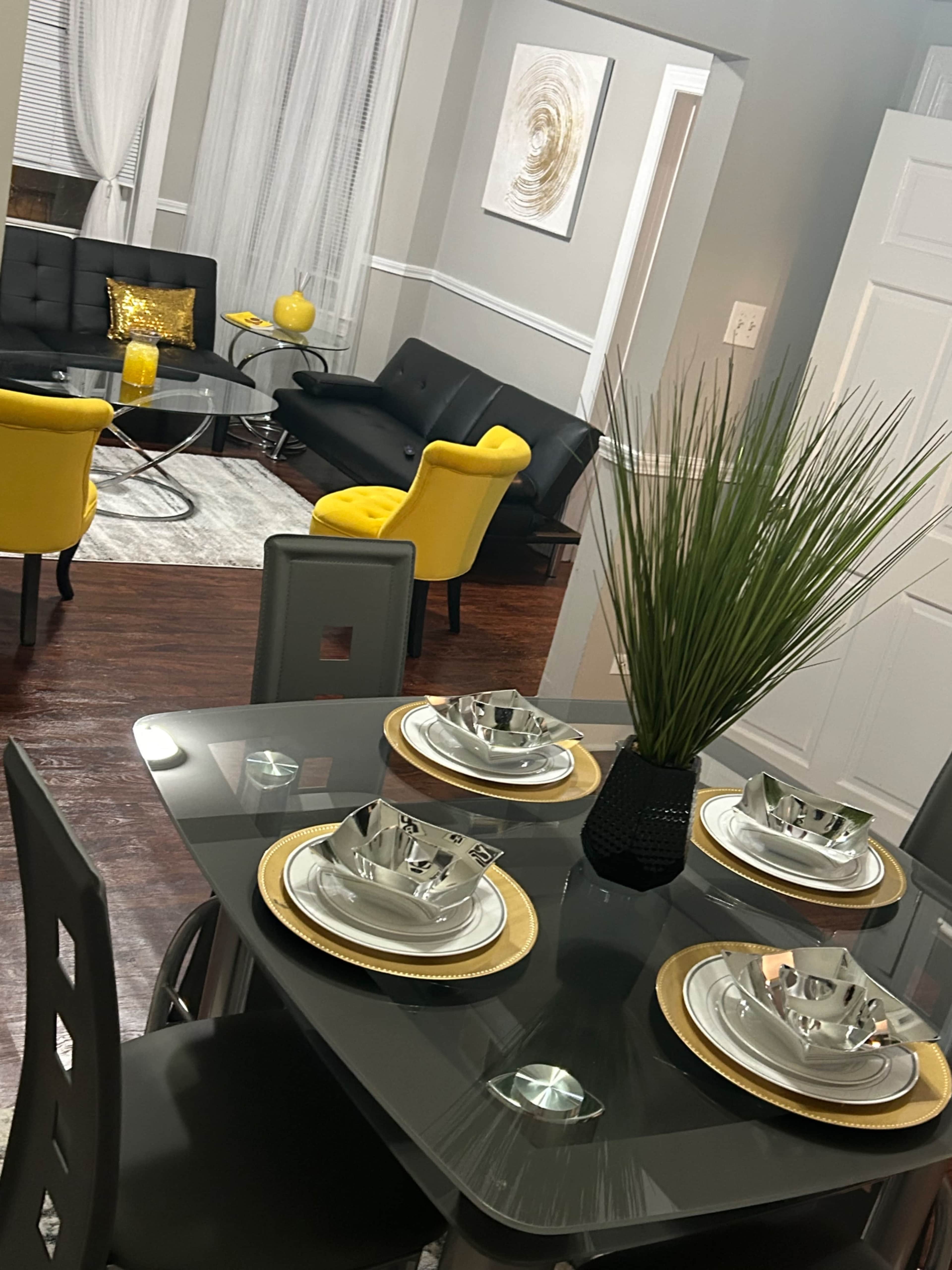 A modern dining area features a glass table set with silver and gold dinnerware, surrounded by sleek chairs, and opens to a stylish living room with yellow accent furniture.