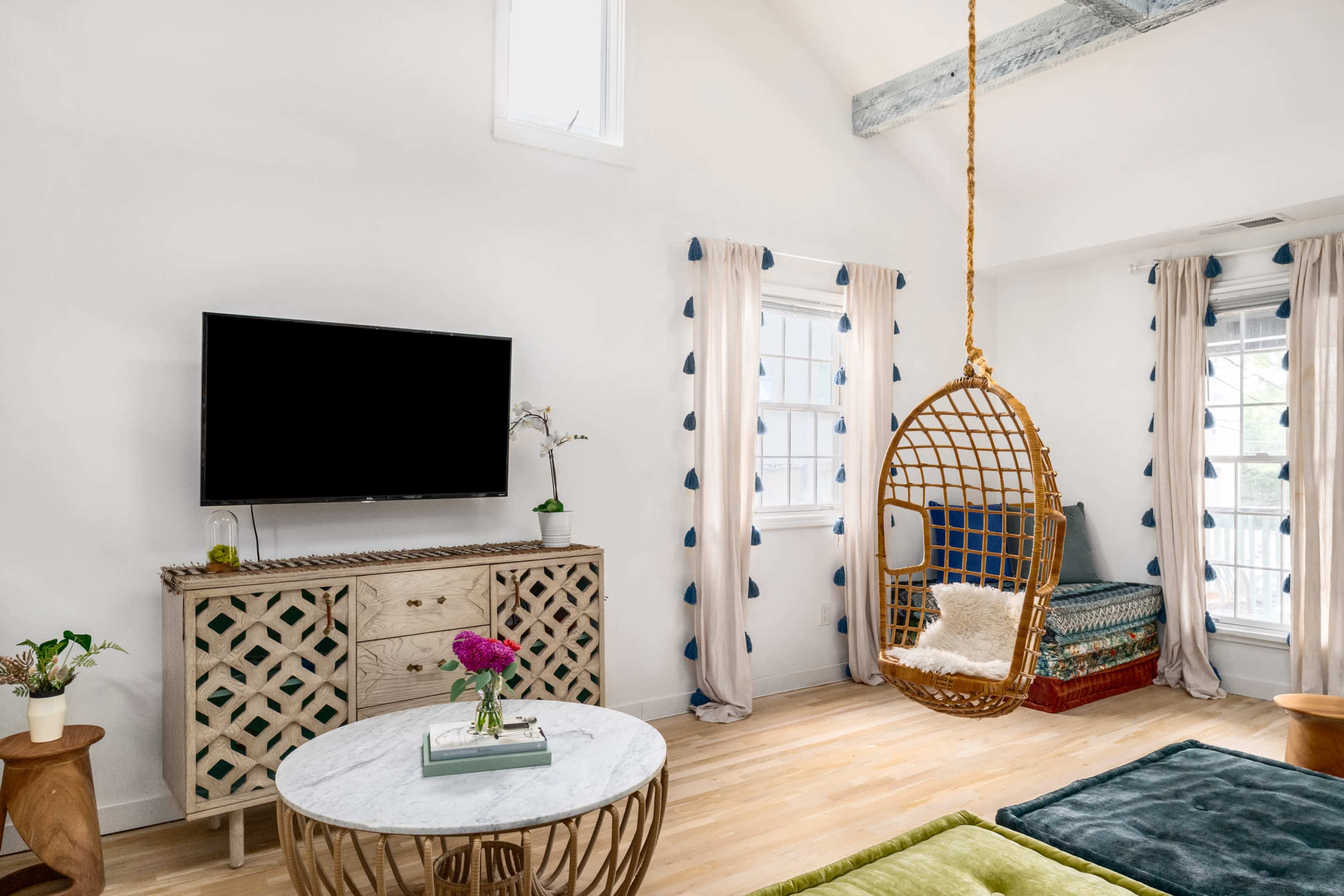 The room features a hanging rattan chair, a television mounted on the wall, and a decorative wooden cabinet, all set against a backdrop of white walls and large windows.