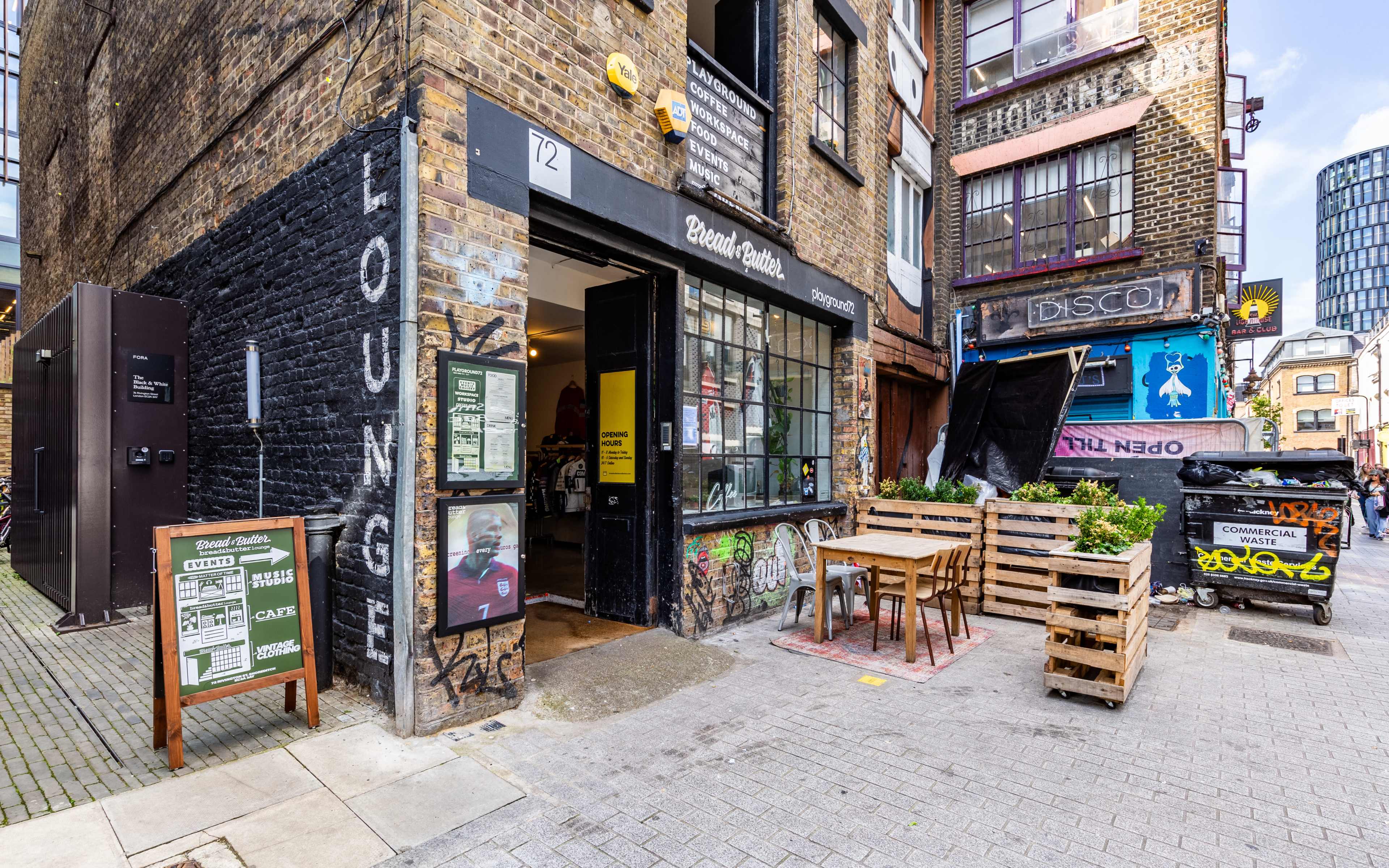 A street view of a brick building entrance labeled "Lounge," with wooden pallets set up as outdoor seating and colorful graffiti on the surrounding walls.
