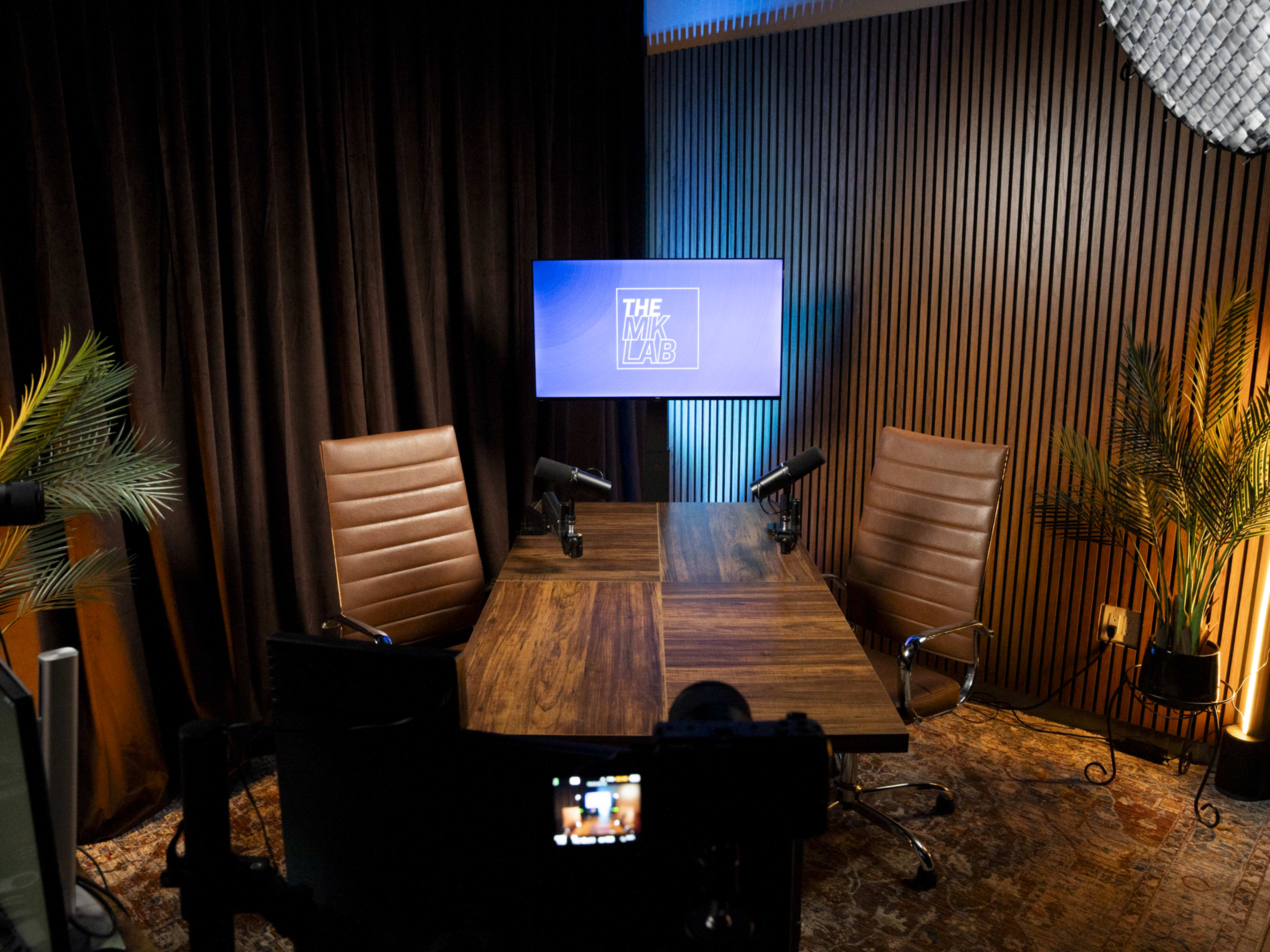 The image shows a modern podcast or video recording studio with two brown leather chairs, a wooden table, and a screen displaying "THE LAB."