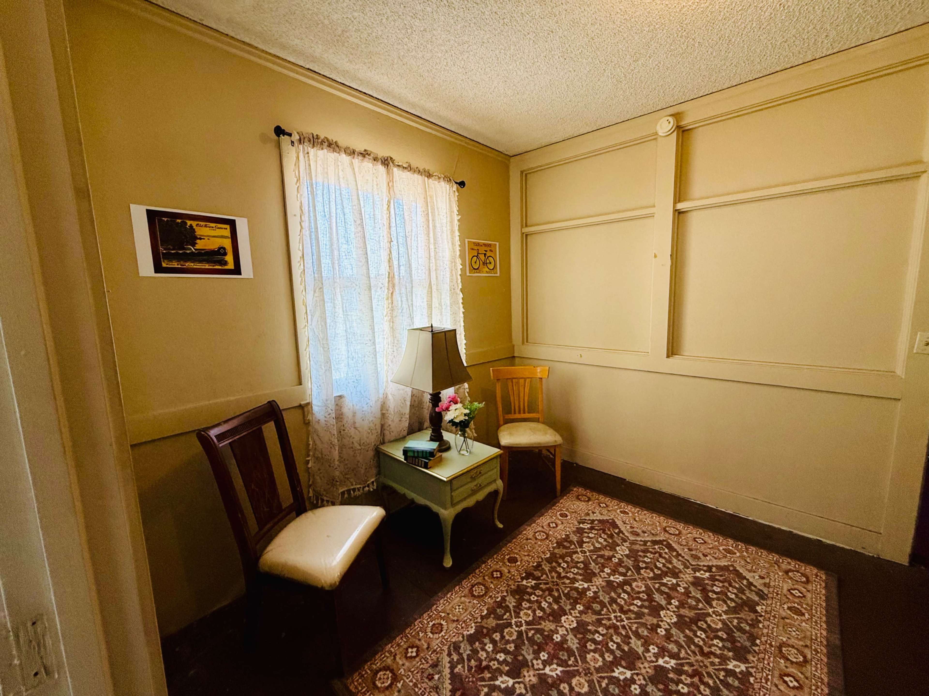 The image shows a small, sparsely furnished room with a window, a table with a vase of flowers, and two chairs.
