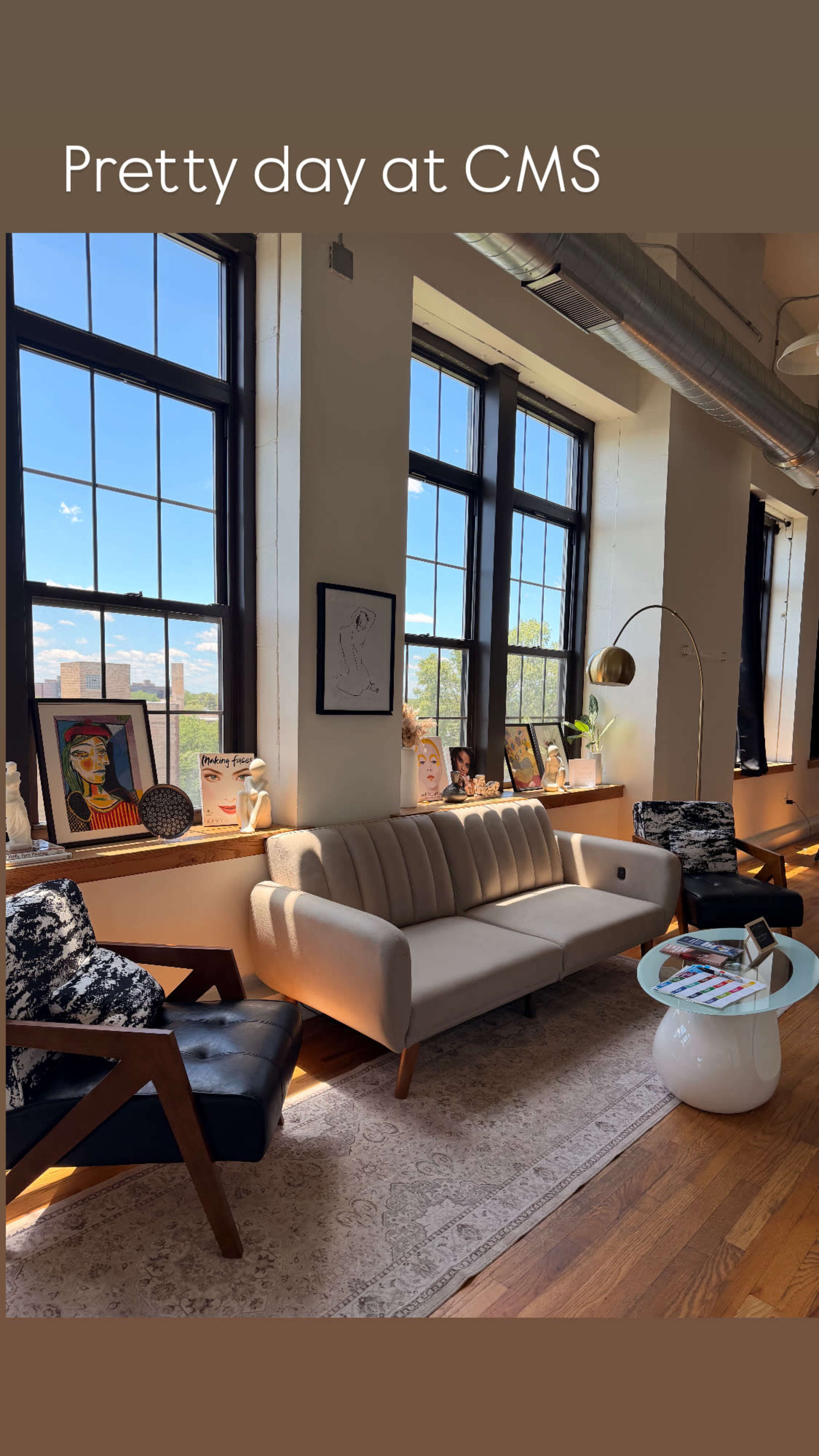 The image shows a bright, open office space with large windows, a white sofa, and chairs, adorned with decorative items and artwork along the walls.