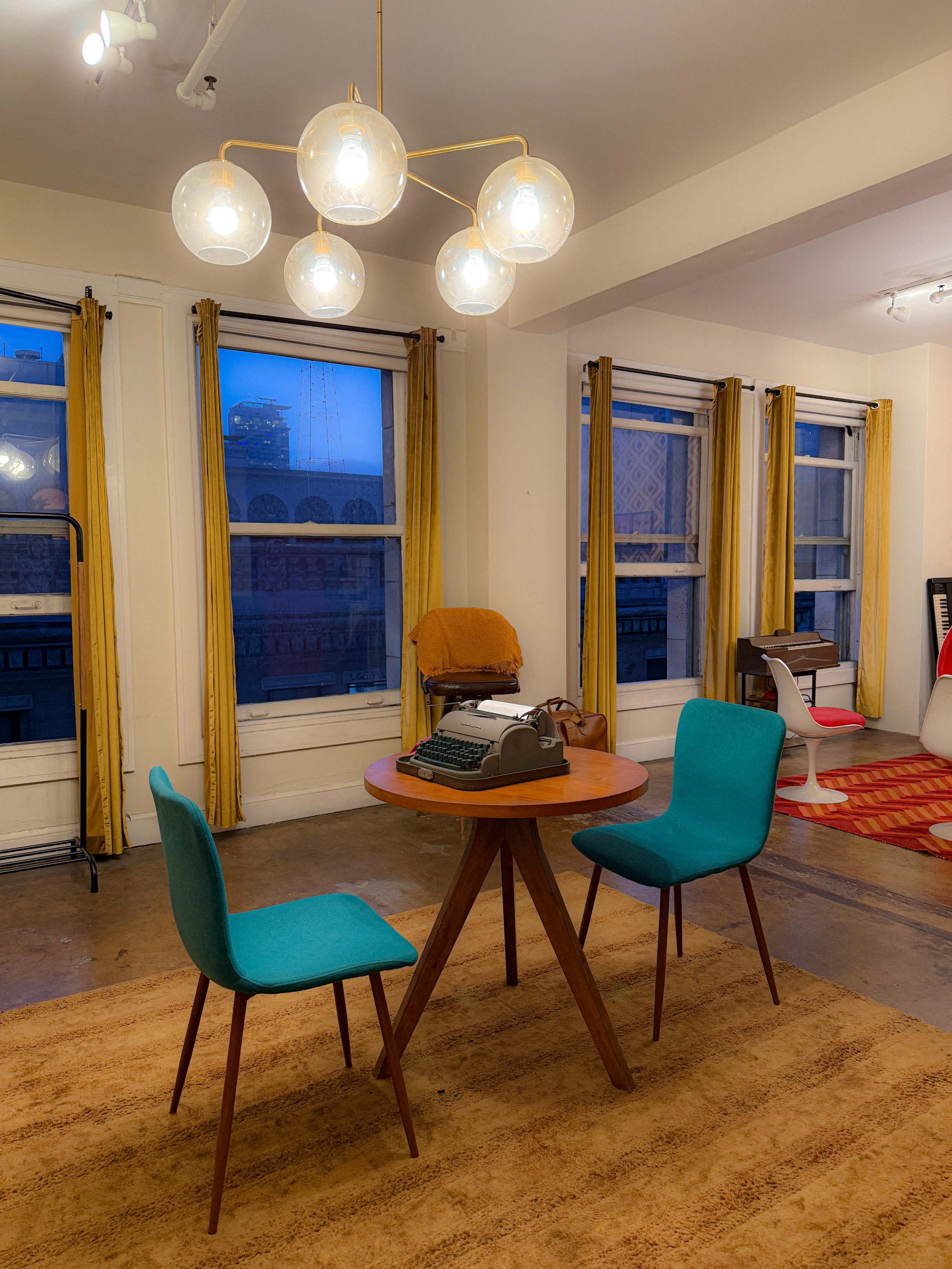 A round wooden table with two teal chairs and a vintage typewriter is set in a room with large windows and yellow curtains.