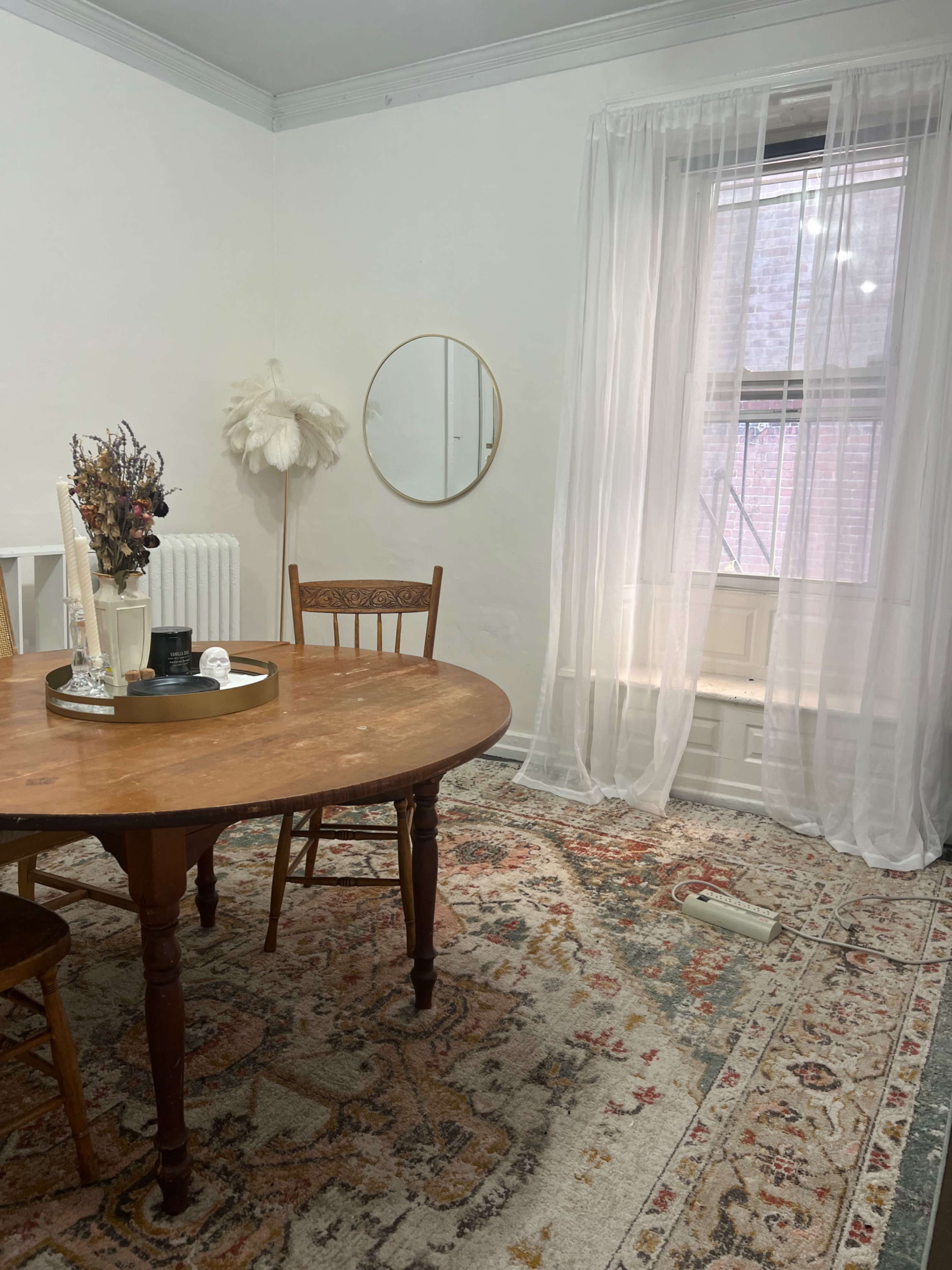 A wooden dining table with chairs is positioned in a room featuring a large window draped with sheer curtains and a round mirror on the wall.