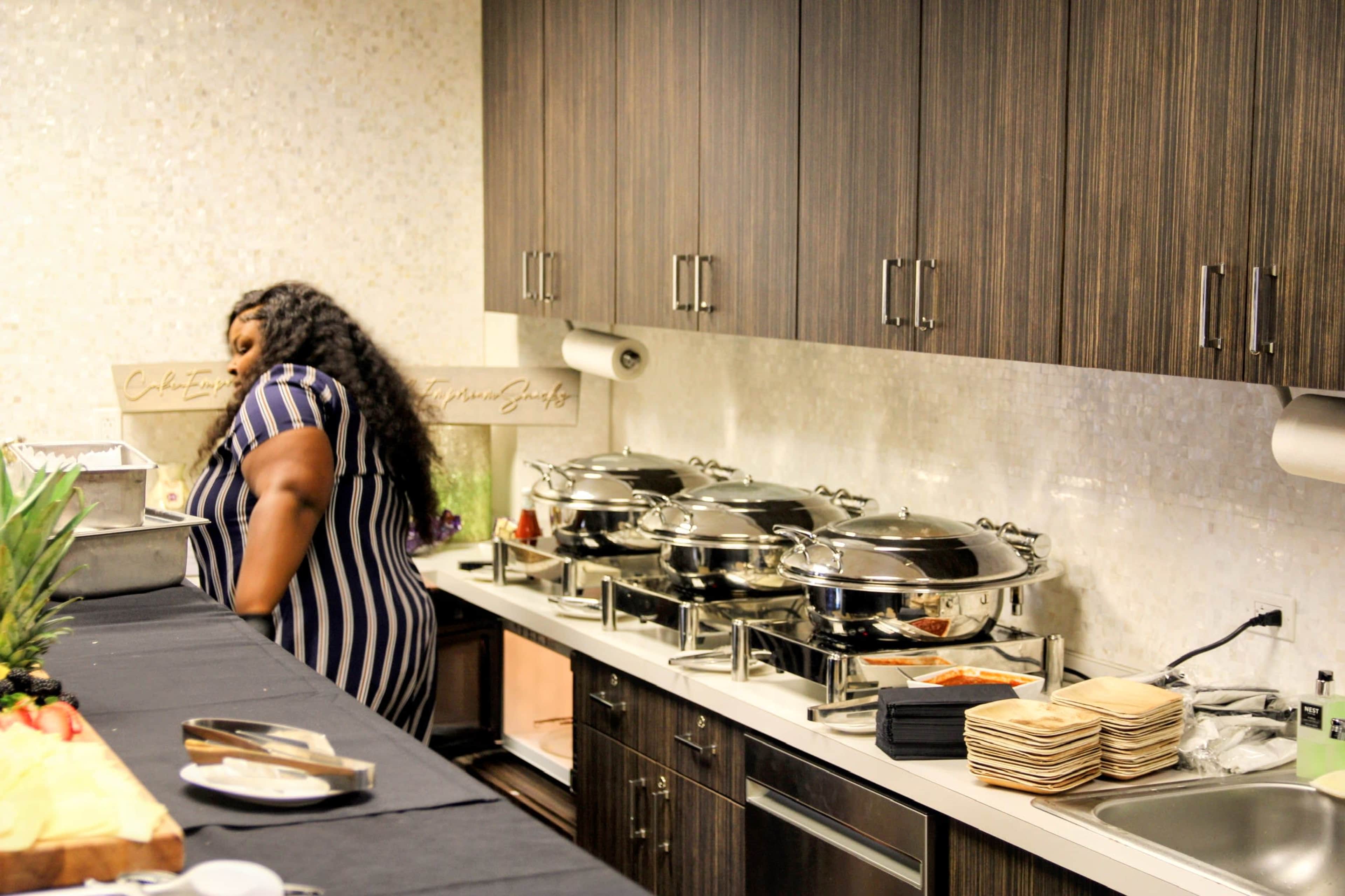 A person in a striped dress stands near a buffet table with three large chafing dishes and stacked plates in a modern kitchen.
