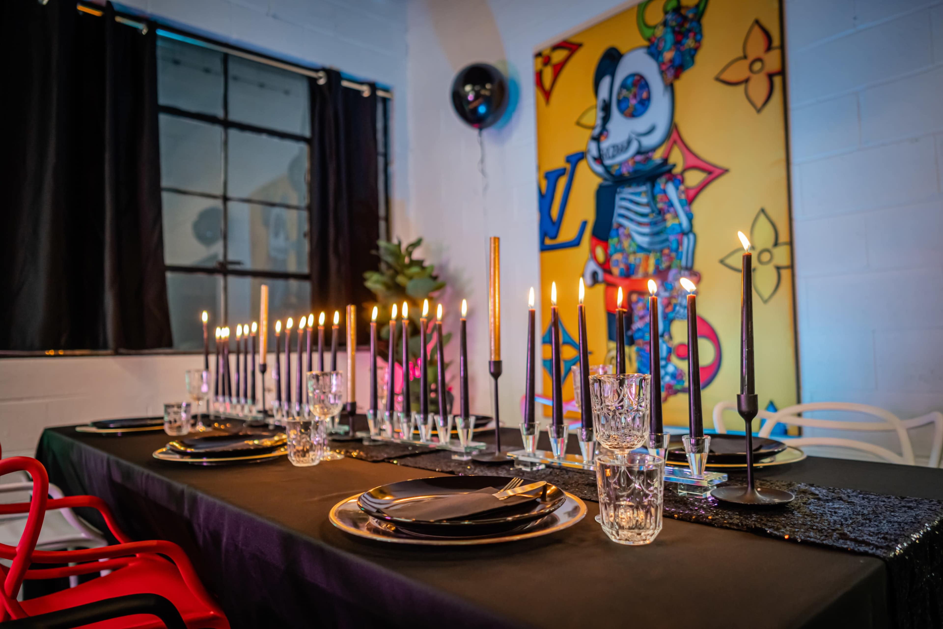 A long black table is set with black plates and glassware, illuminated by numerous candlesticks, against a backdrop featuring a colorful artwork and black curtains.