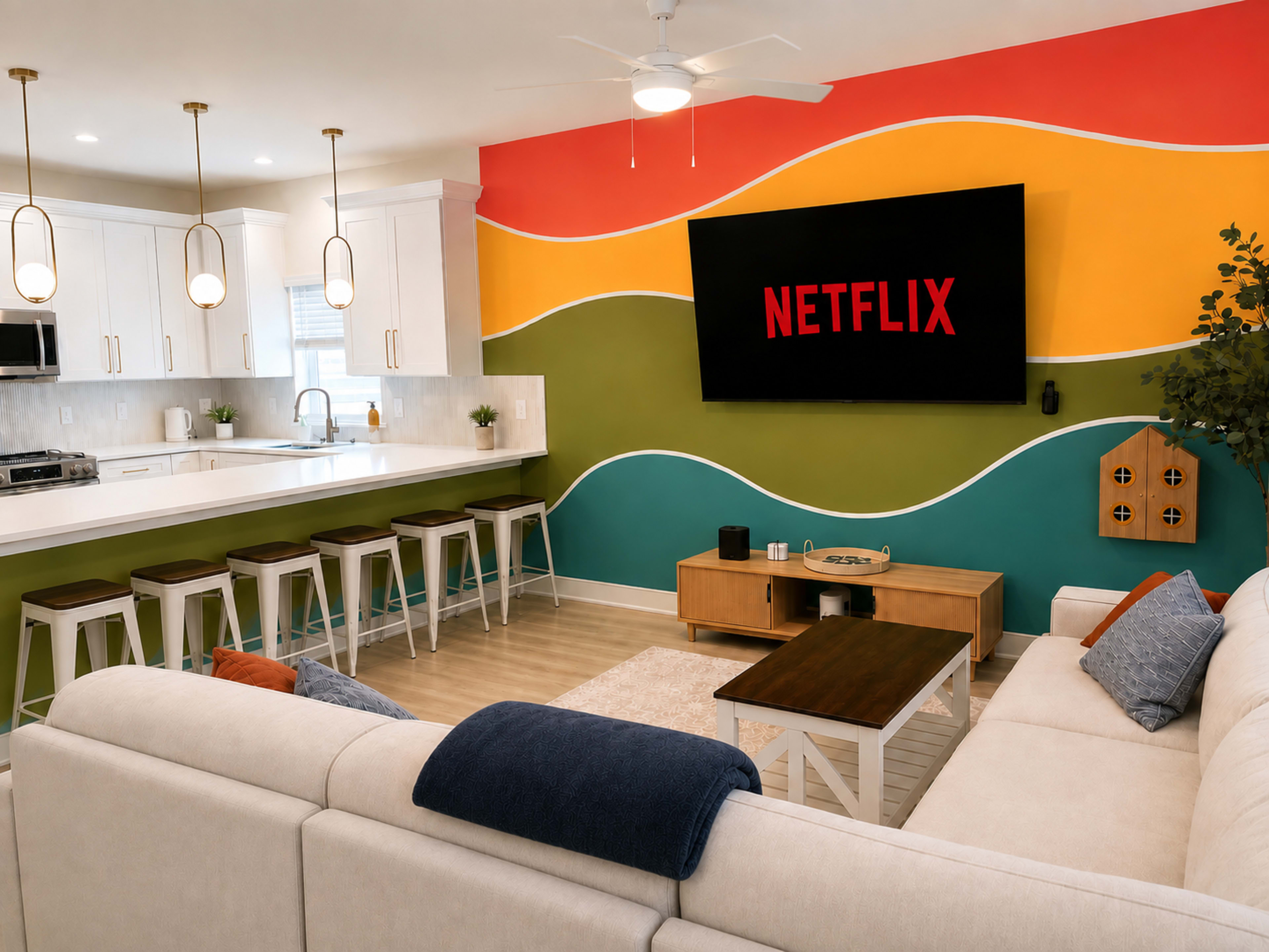 The image shows a modern living room featuring a large wall-mounted television displaying the Netflix logo, a beige sectional sofa, a wooden coffee table, white bar stools at a kitchen island, and a colorful painted accent wall.