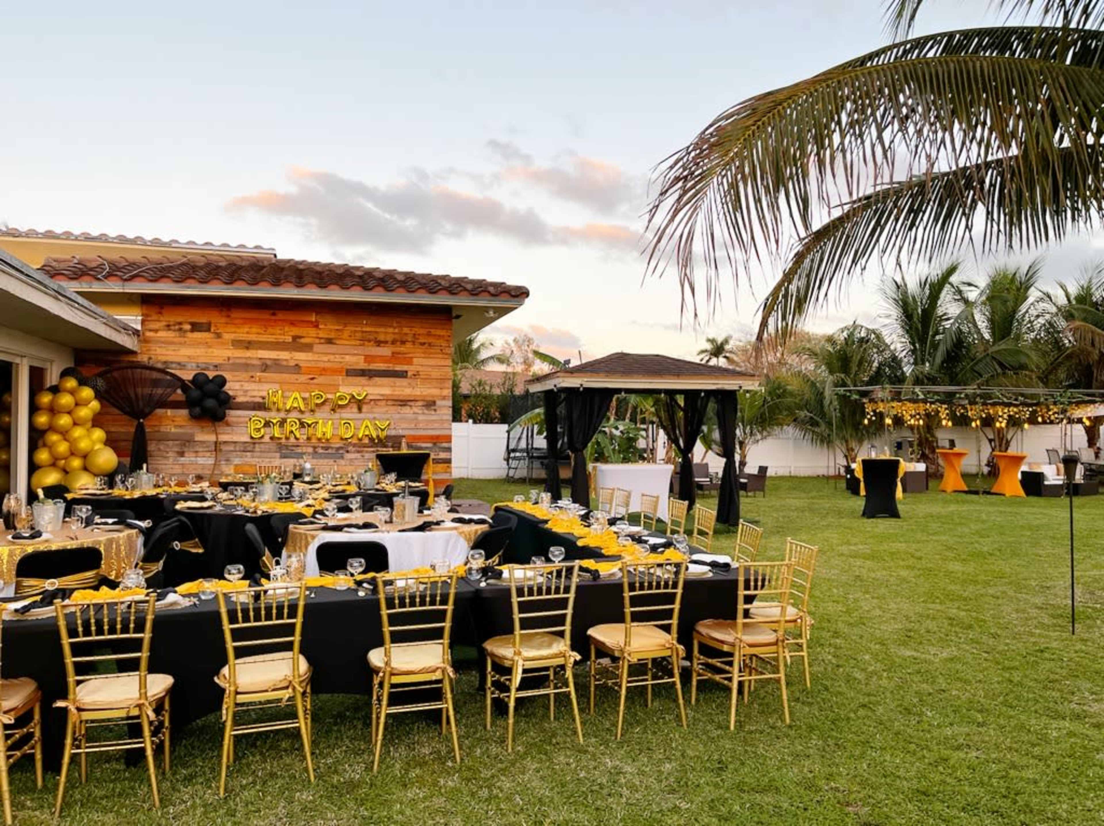 An outdoor birthday party setup features a decorated table with gold and black accents, surrounded by chairs, in a yard with palm trees and a gazebo.