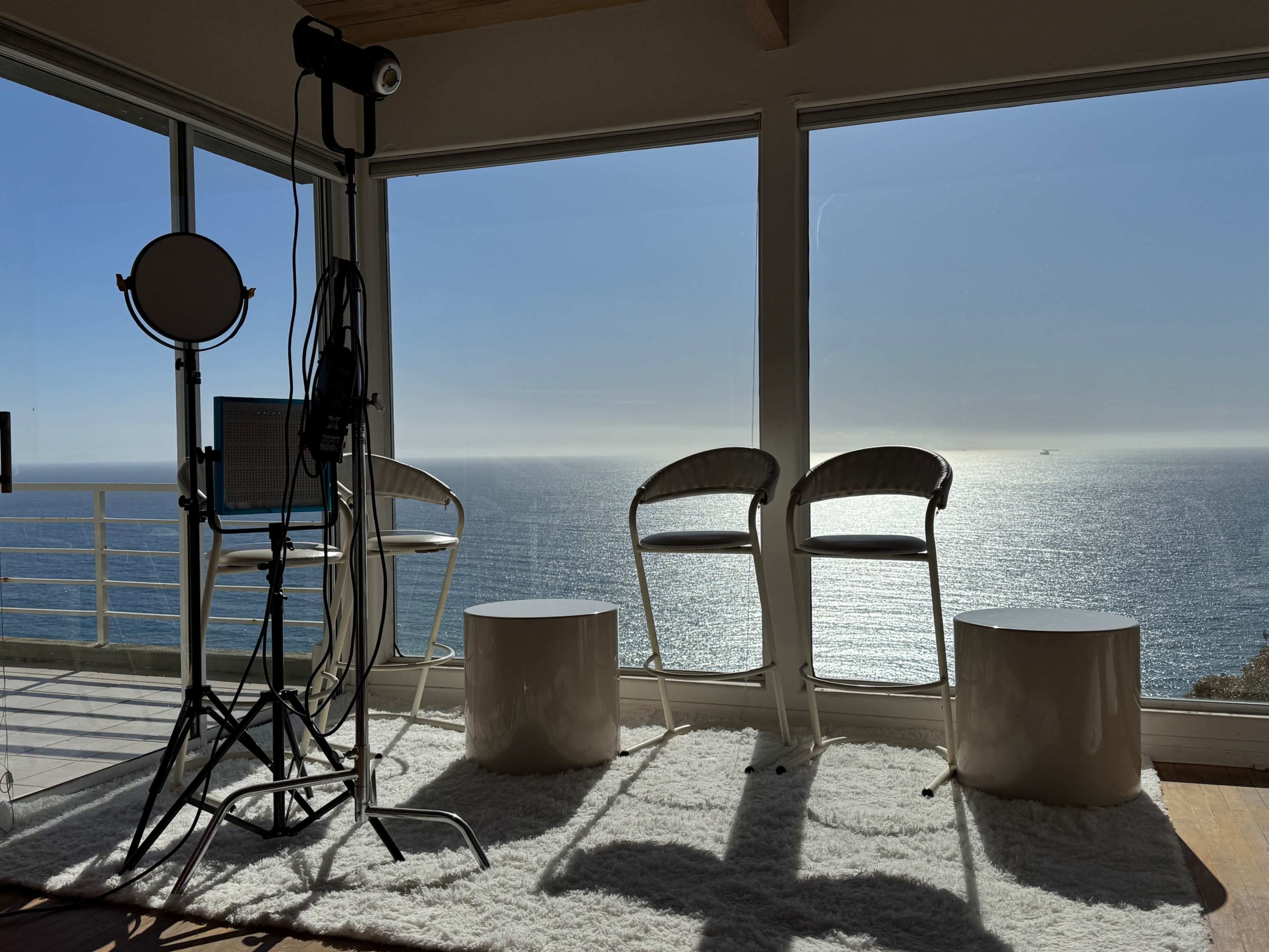 The image shows a bright room with three chairs and two small tables facing a large window overlooking the ocean.