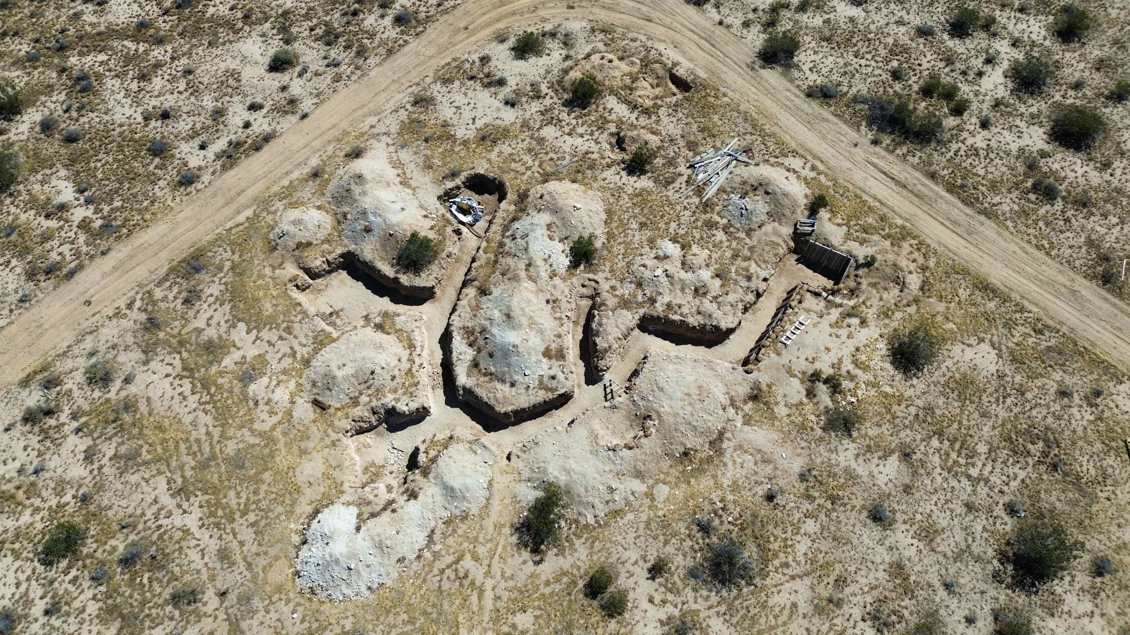 The image shows an aerial view of a barren landscape with several earth mounds and trenches arranged in a winding pattern near a dirt road.