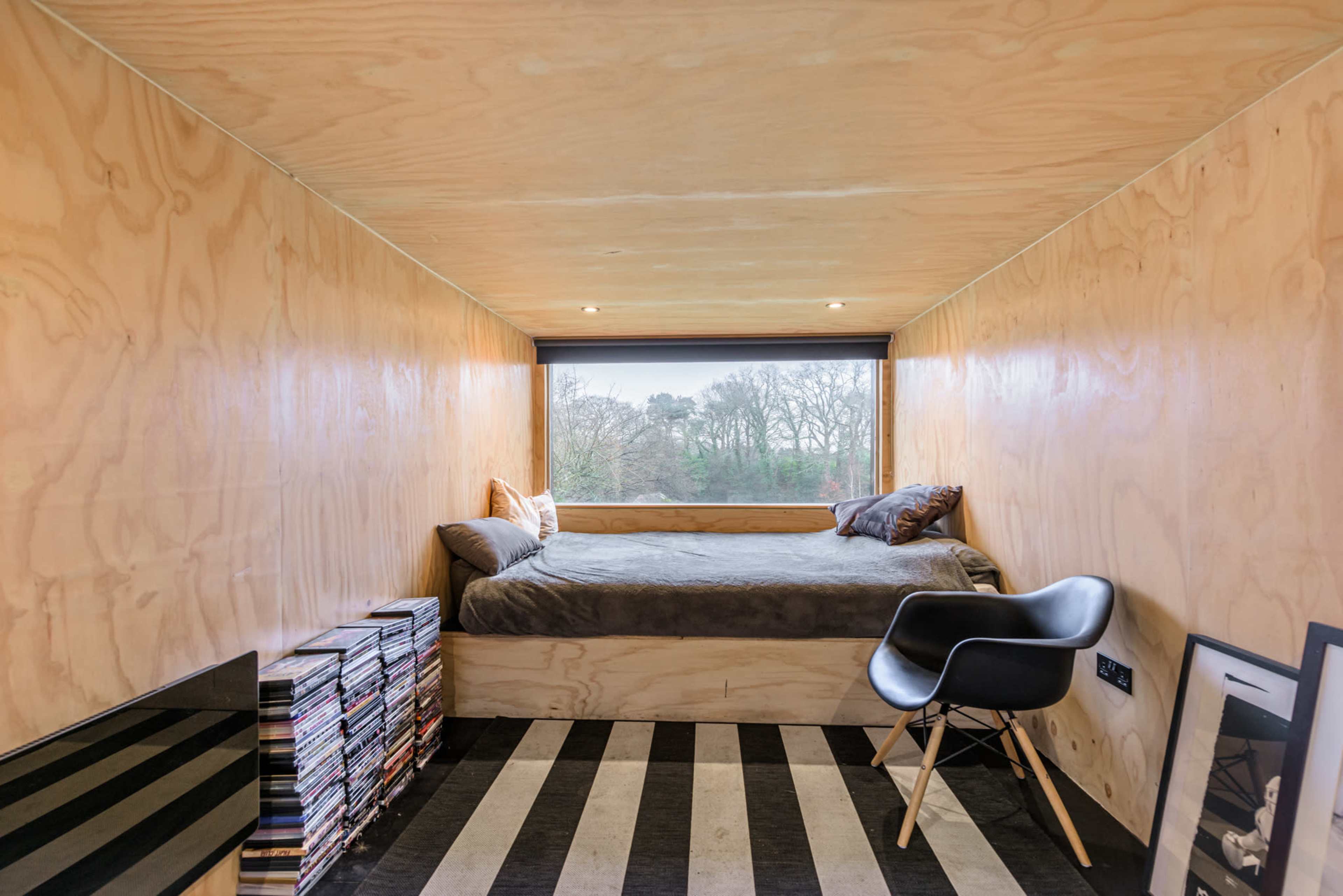 The image shows a minimalist bedroom with a bed, a stack of DVDs, and a large window overlooking a tree-filled landscape.