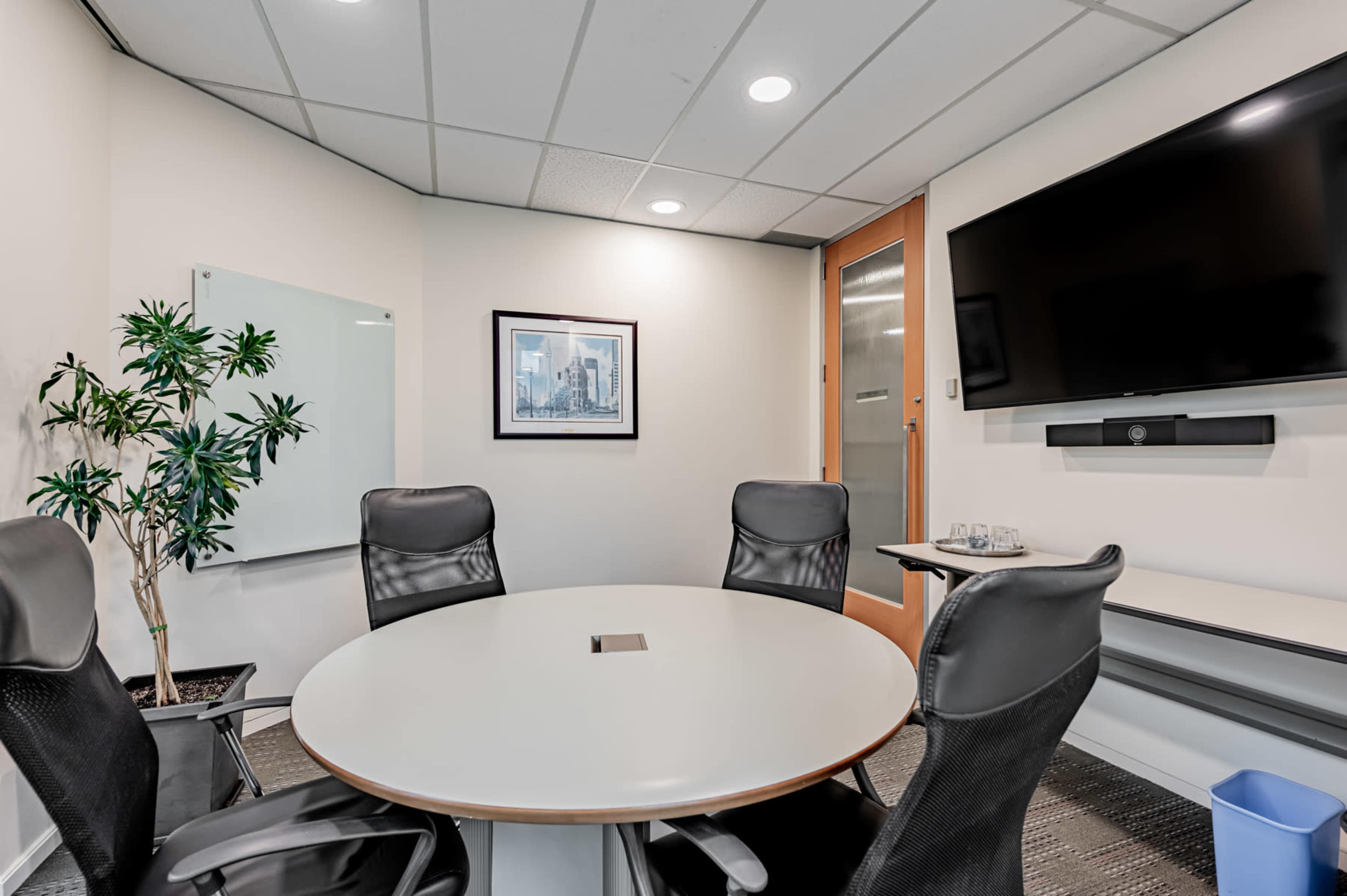 A modern conference room features a round table surrounded by black ergonomic chairs, a large wall-mounted television, and a glass door.
