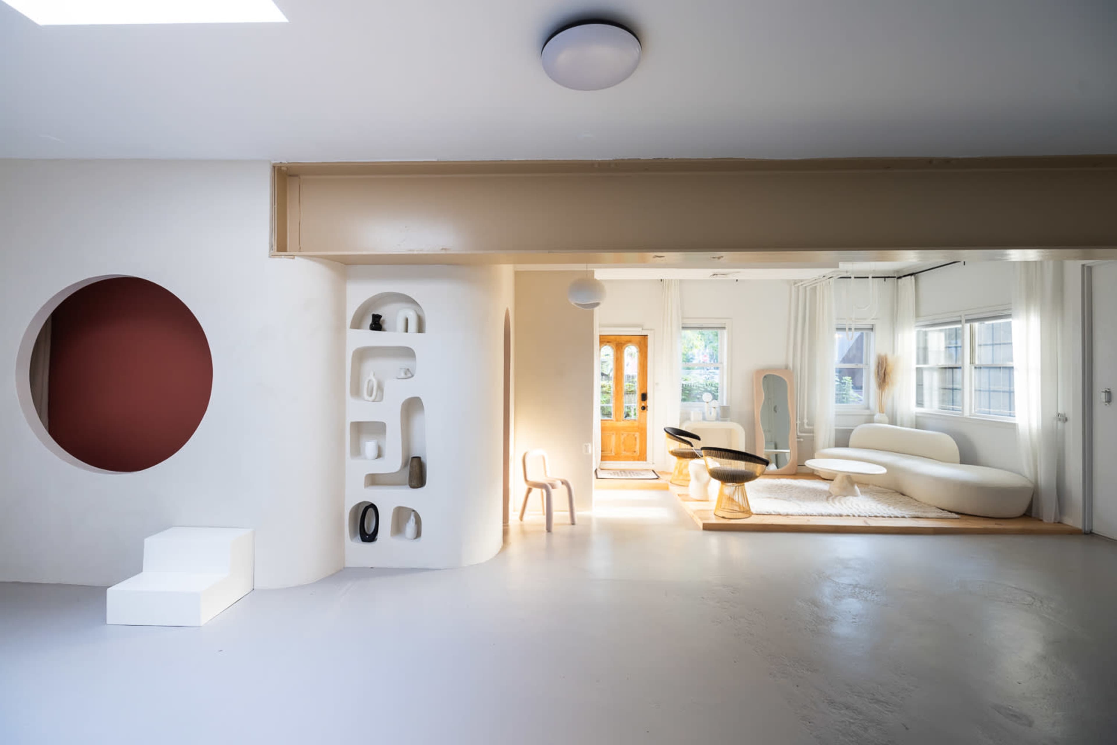 A spacious interior features a circular opening, sculptural wall shelves, a minimalist staircase, and modern furniture arranged around large windows.