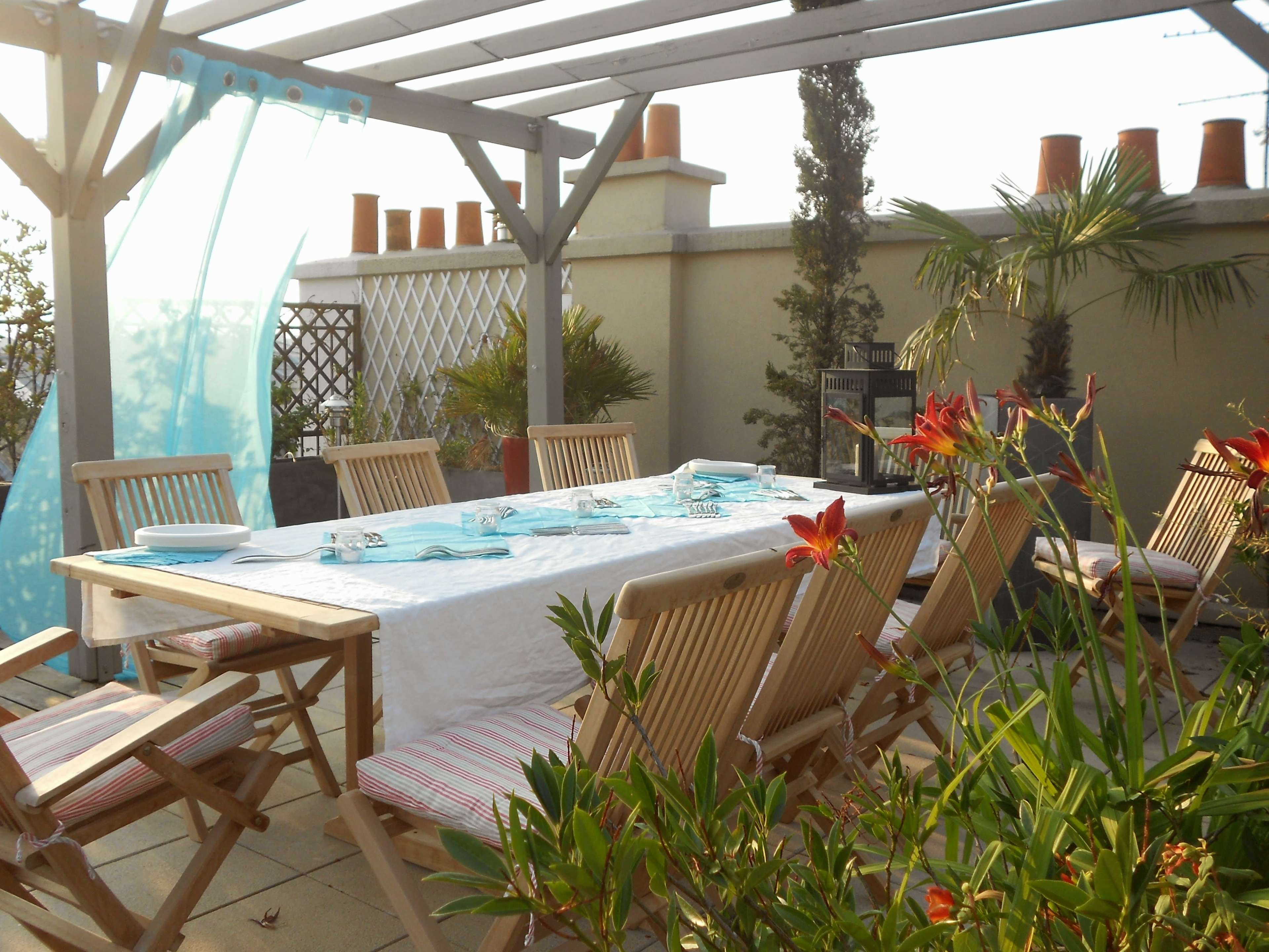 A wooden dining table is set with blue glassware and surrounded by chairs on a terrace adorned with potted plants.