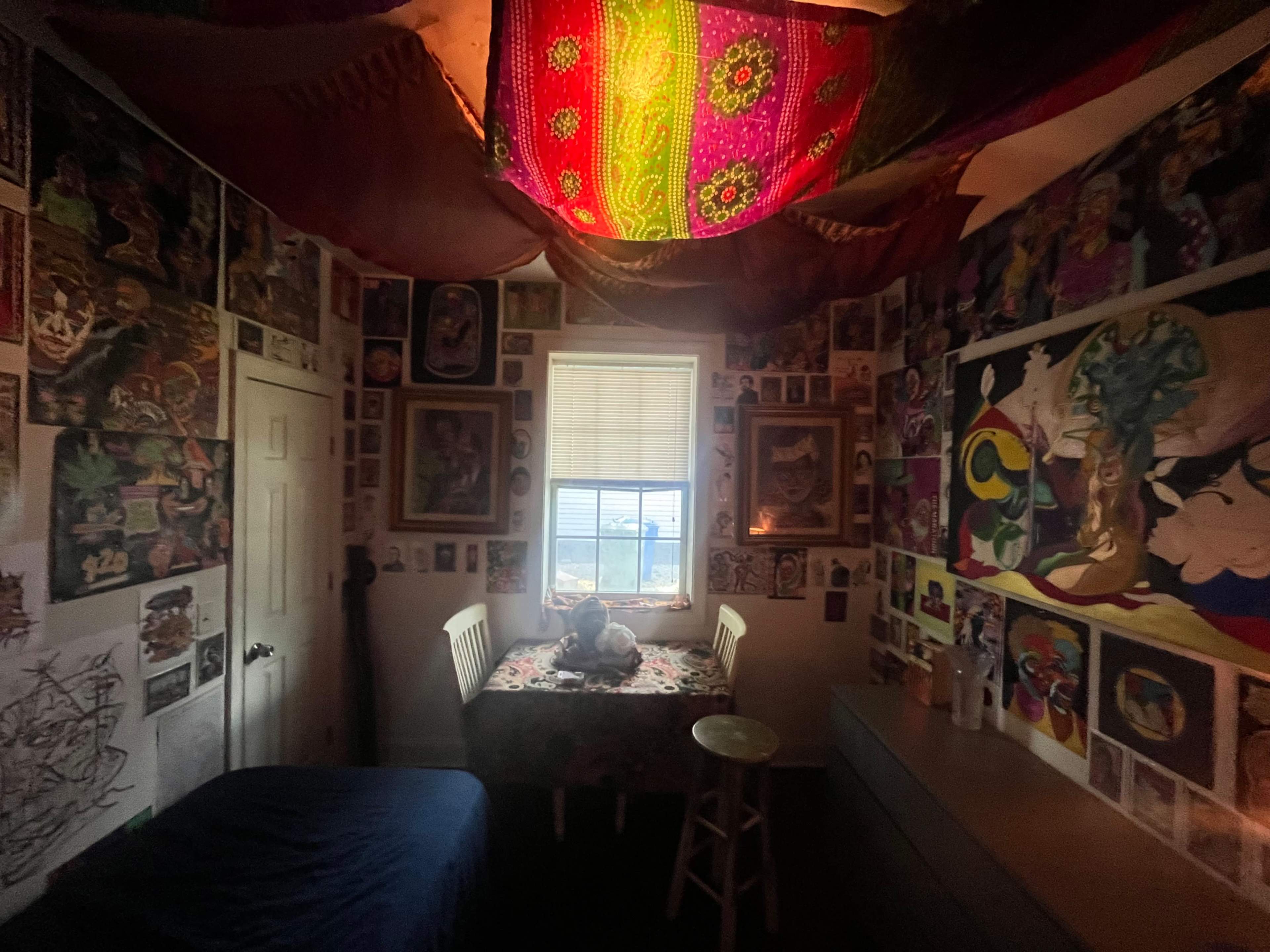 The room features a table with a stool, a bed, and walls covered with various artworks and posters, all under a colorful fabric ceiling.