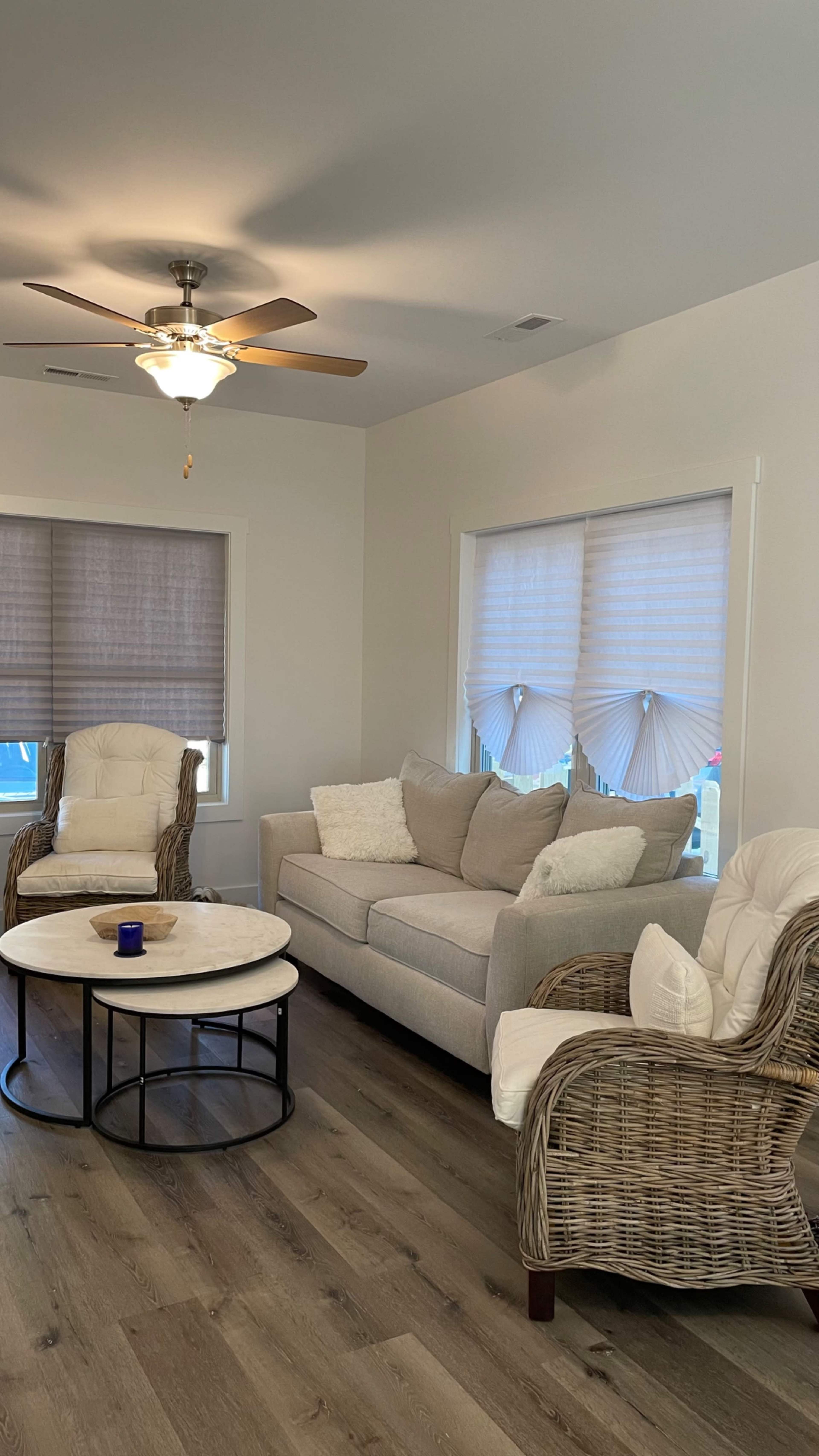 The image shows a living room with a gray sofa, two wicker chairs, a round coffee table, and windows featuring pleated shades.