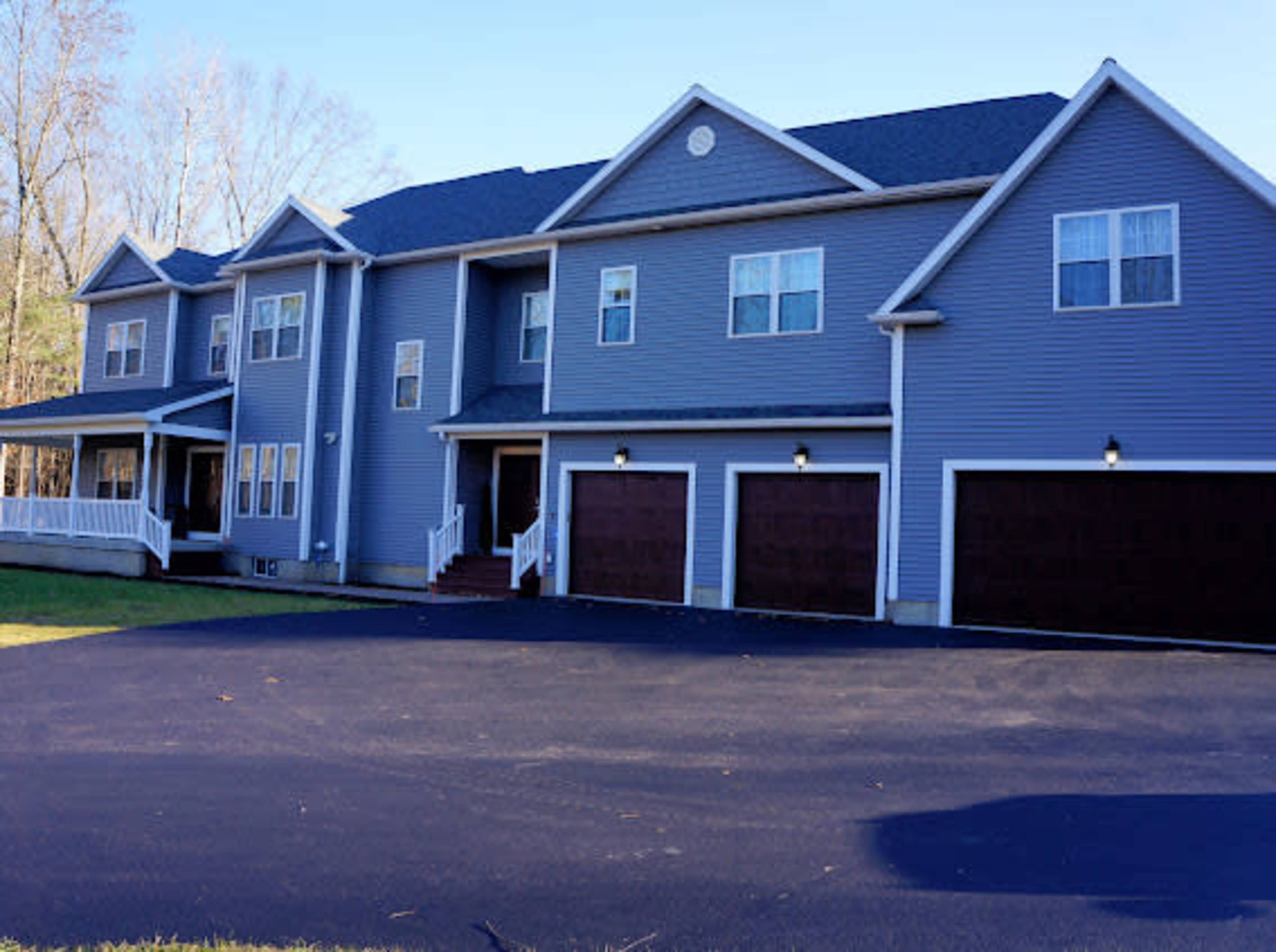 A two-story blue house with a three-car garage is set on a paved driveway surrounded by trees.