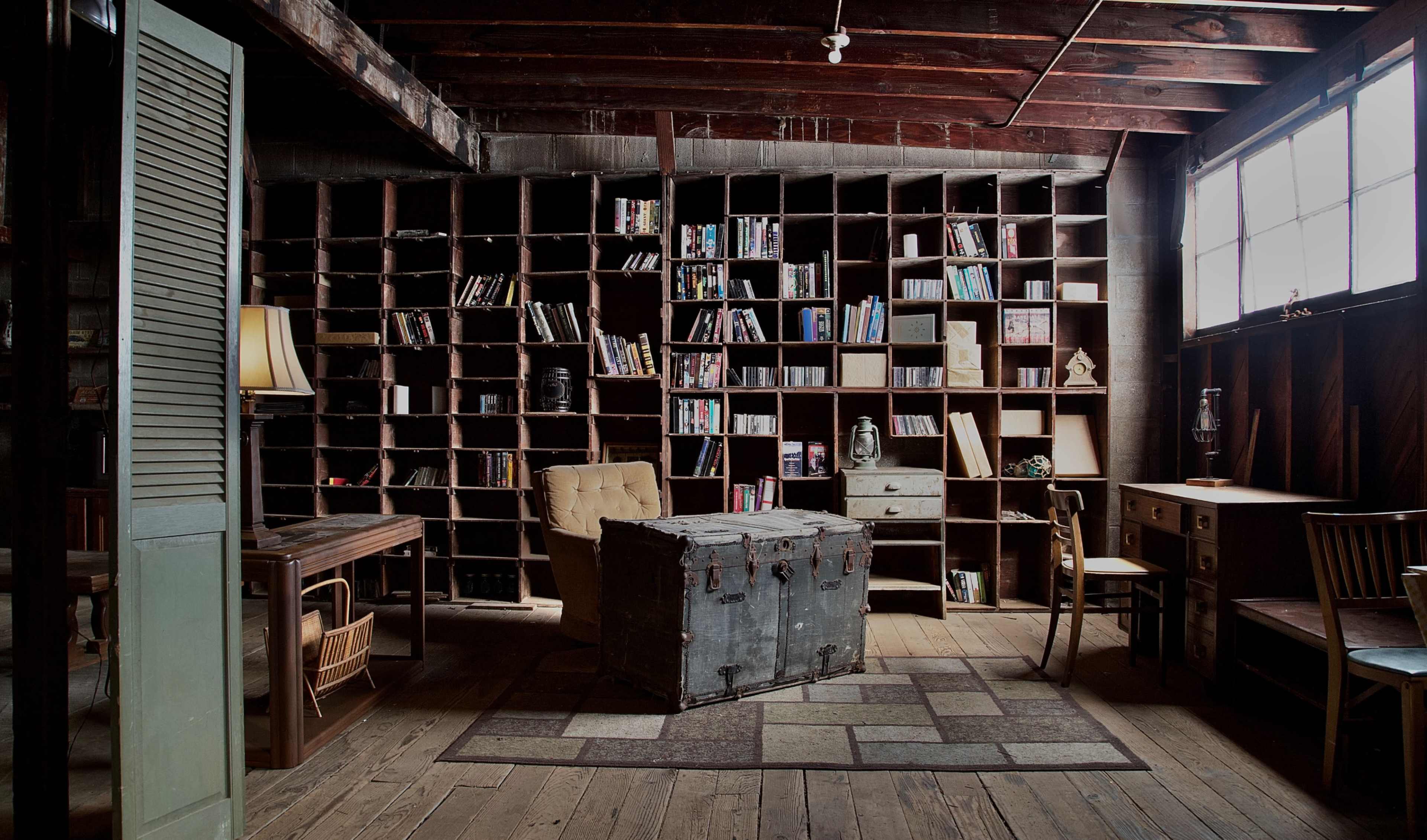 A cozy library with wooden shelving filled with books and a vintage trunk in the center of the room alongside a desk and chairs.