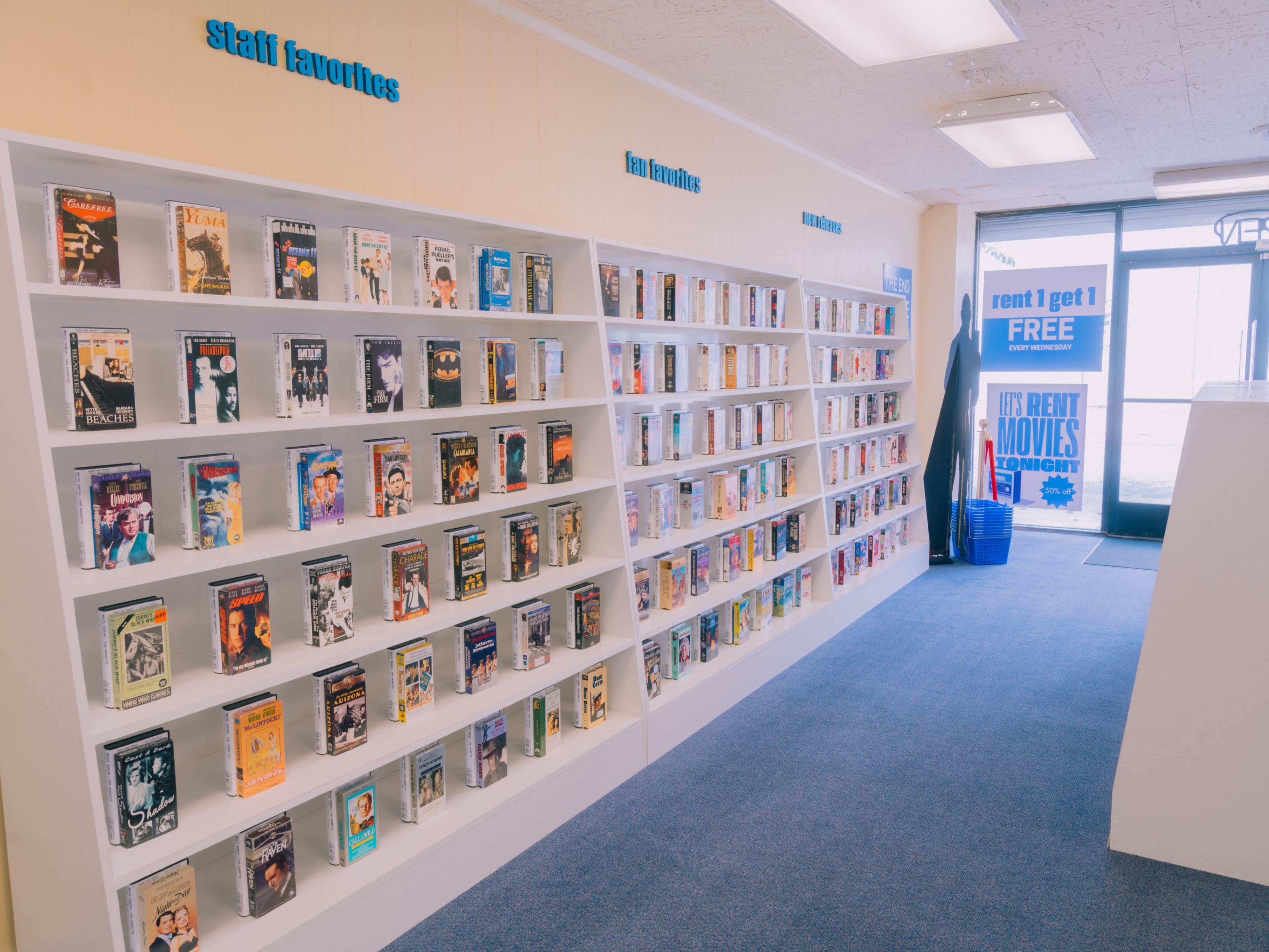The image shows an interior of a video rental store with two walls lined with shelves containing various VHS tapes.