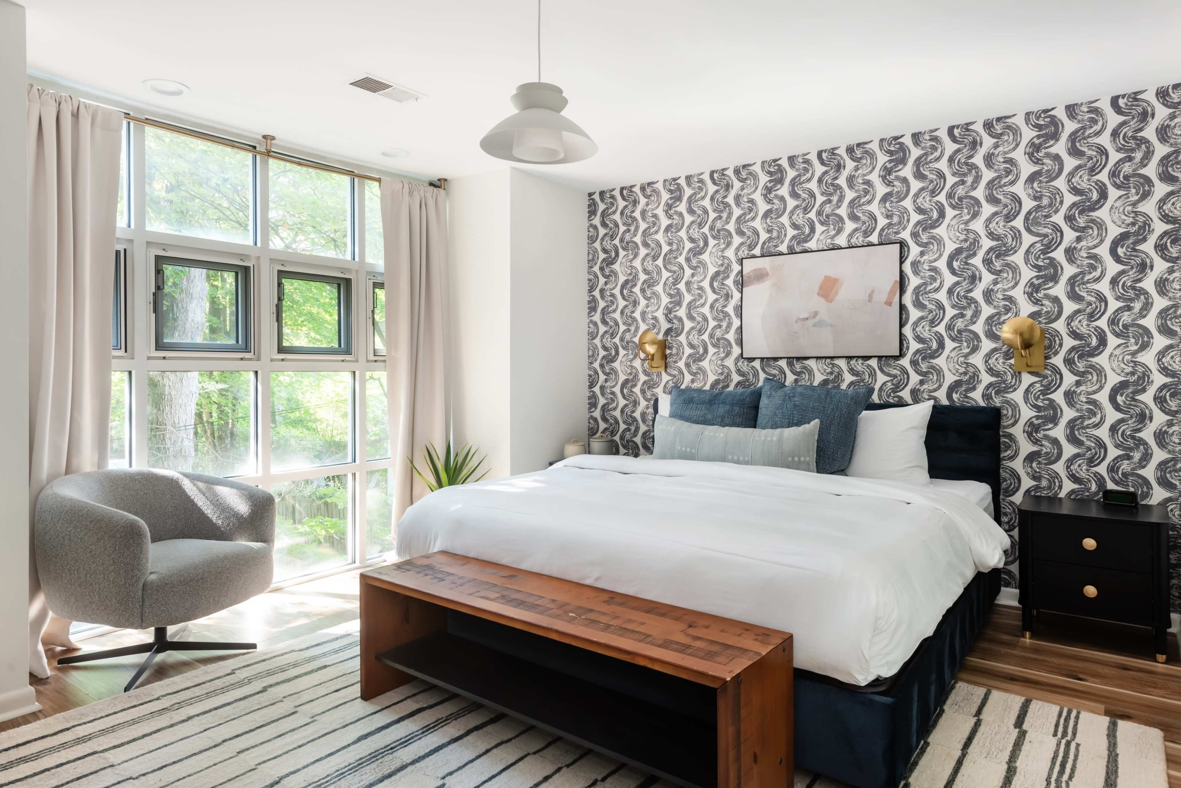 The image shows a stylish bedroom featuring a bed with blue and white bedding, a wooden bench at the foot, a gray armchair, and large windows that provide natural light, all against a patterned wallpaper.