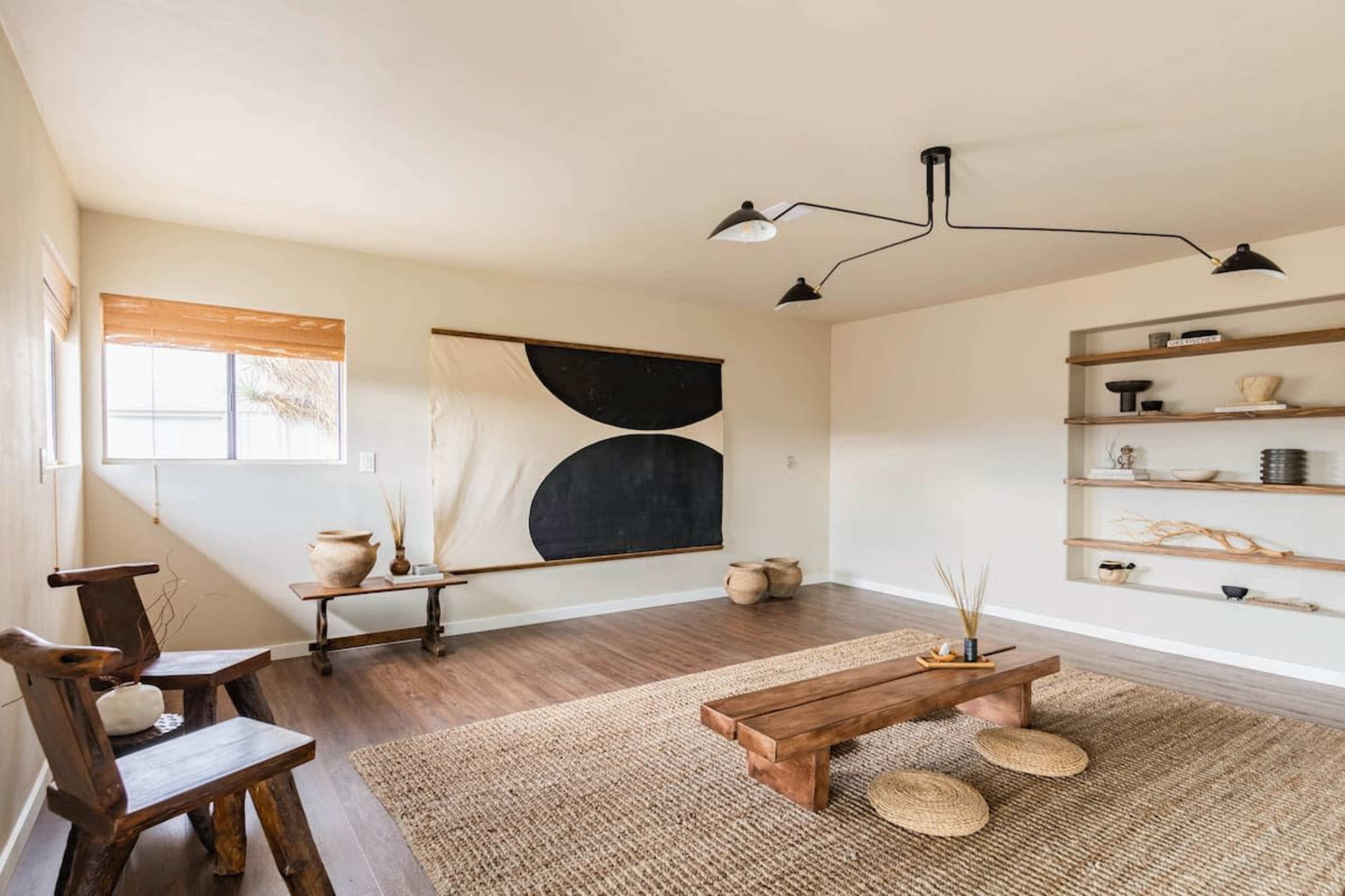 A minimalist living room features a low wooden coffee table, a woven rug, a wall art piece with black and beige shapes, and wooden shelves displaying decorative items.