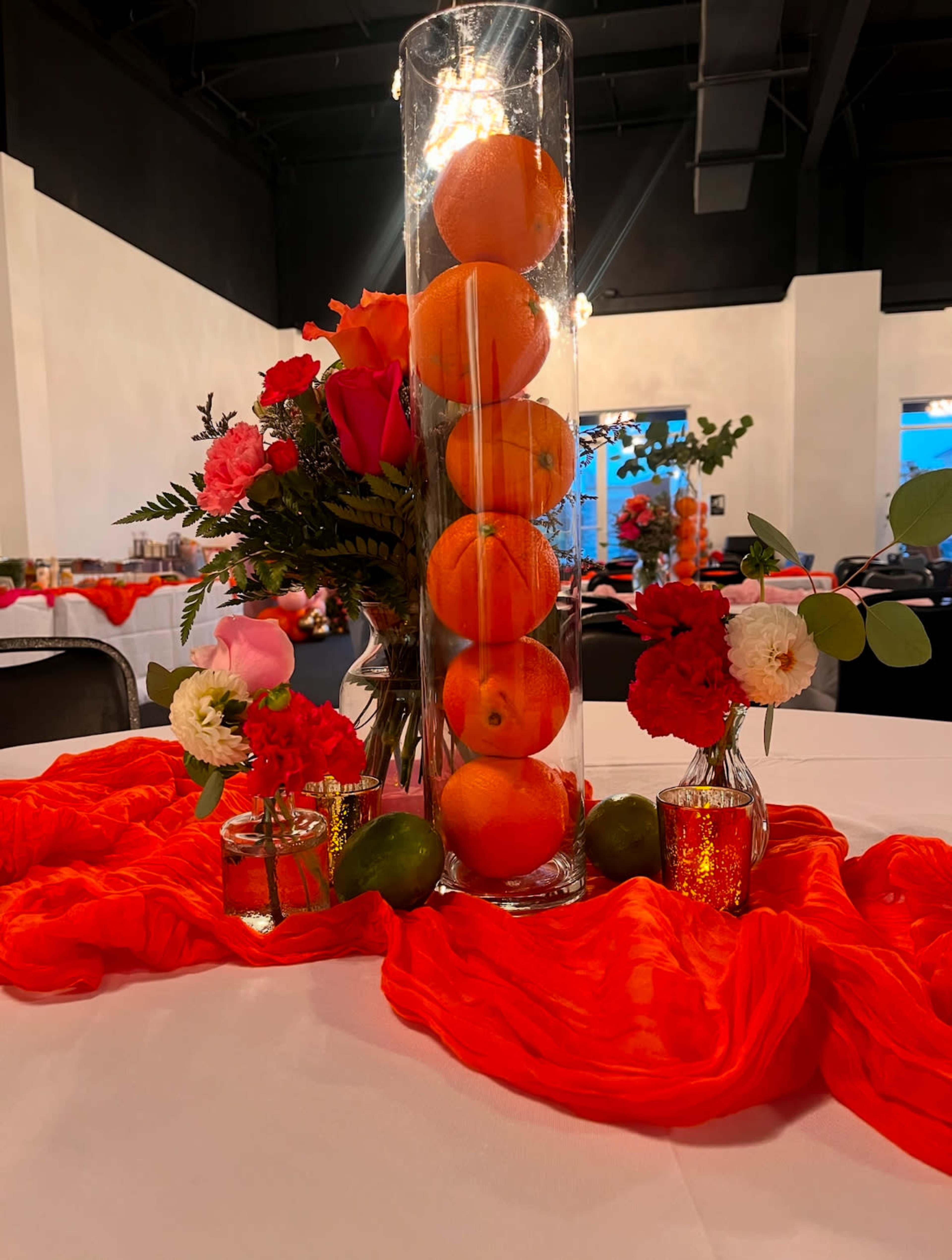 A centerpiece features a tall glass cylinder filled with oranges, surrounded by small vases of colorful flowers on a table draped with a vibrant orange cloth.