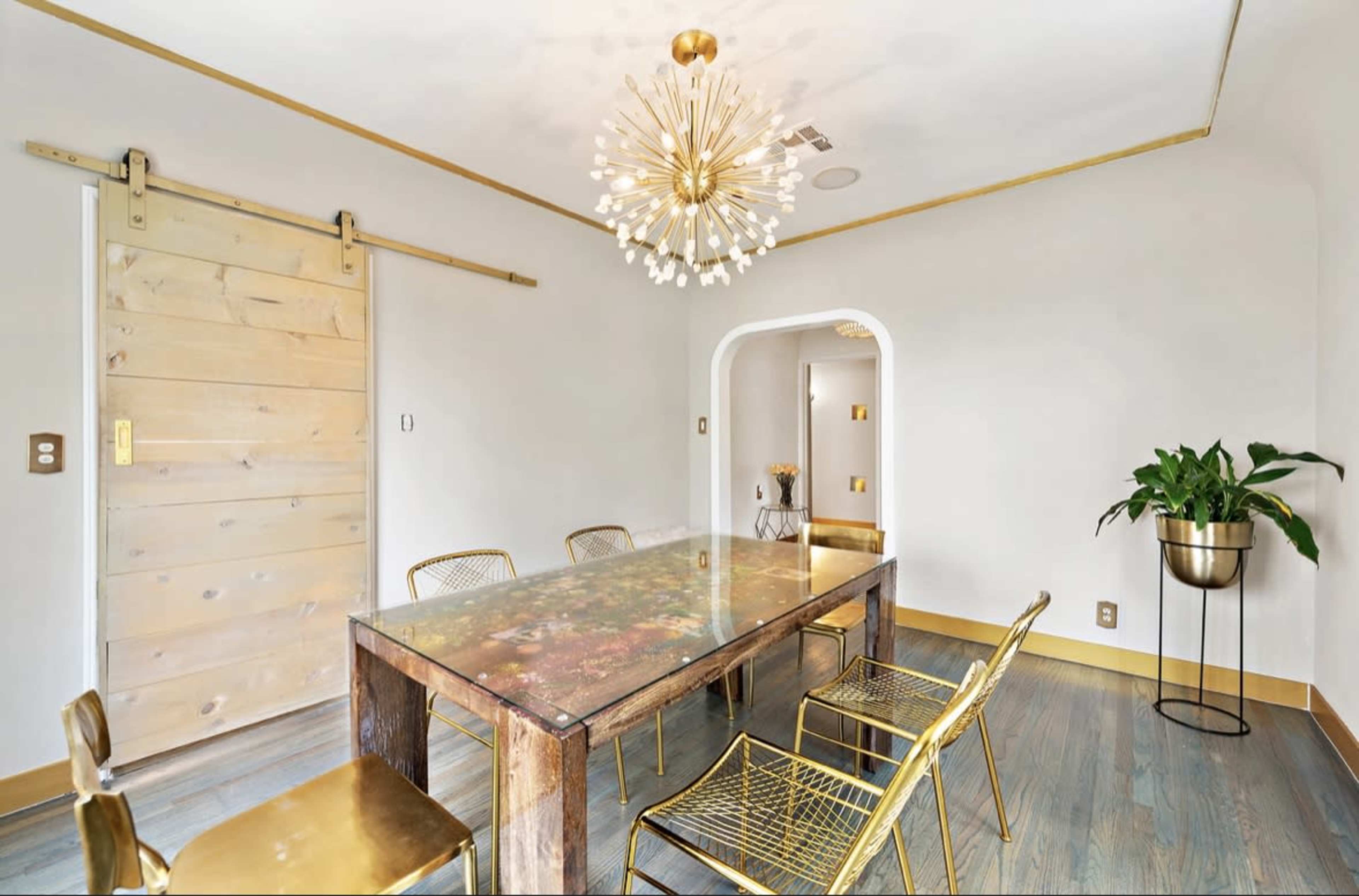 A dining room features a floral-patterned table with six gold metal chairs, a chandelier, and a sliding wooden door.