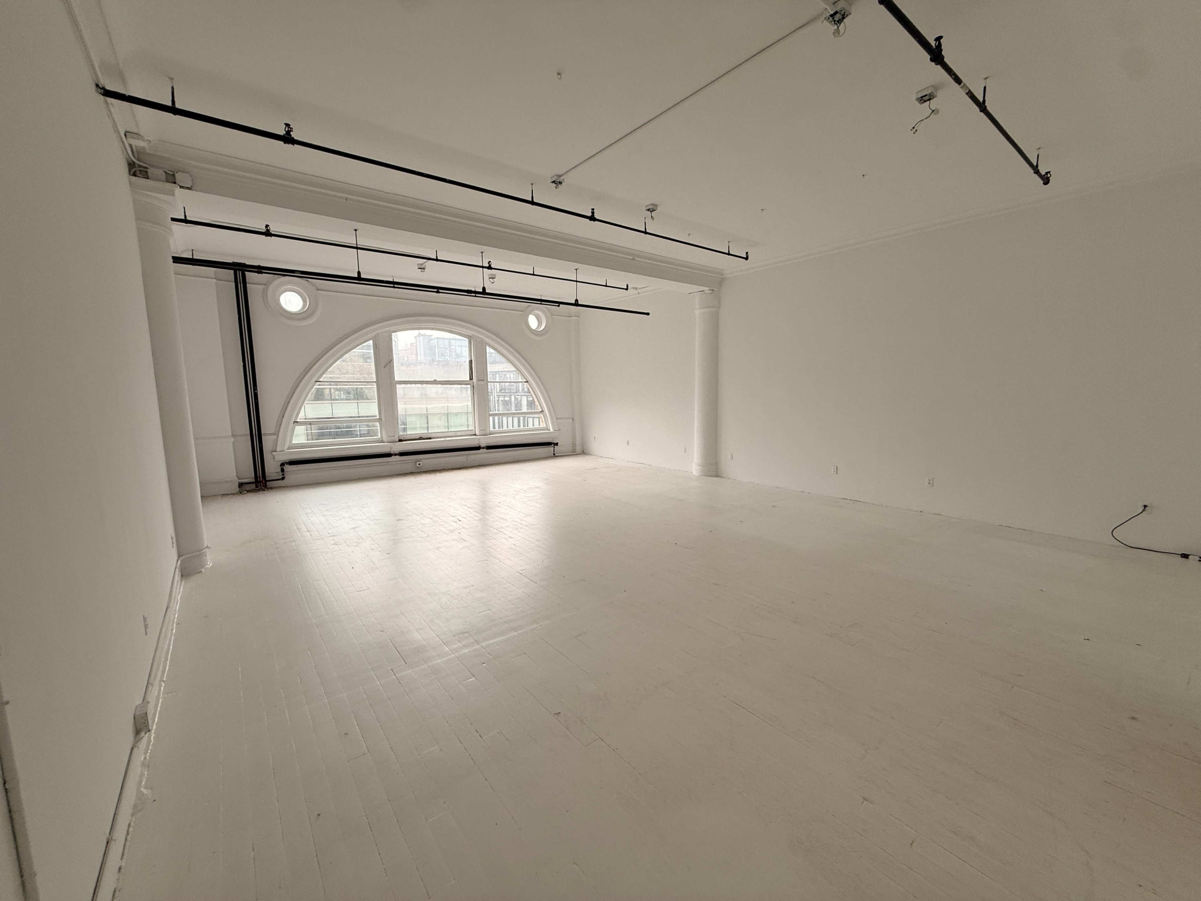 The image shows an empty, white-walled room with large arched windows and a polished wooden floor.