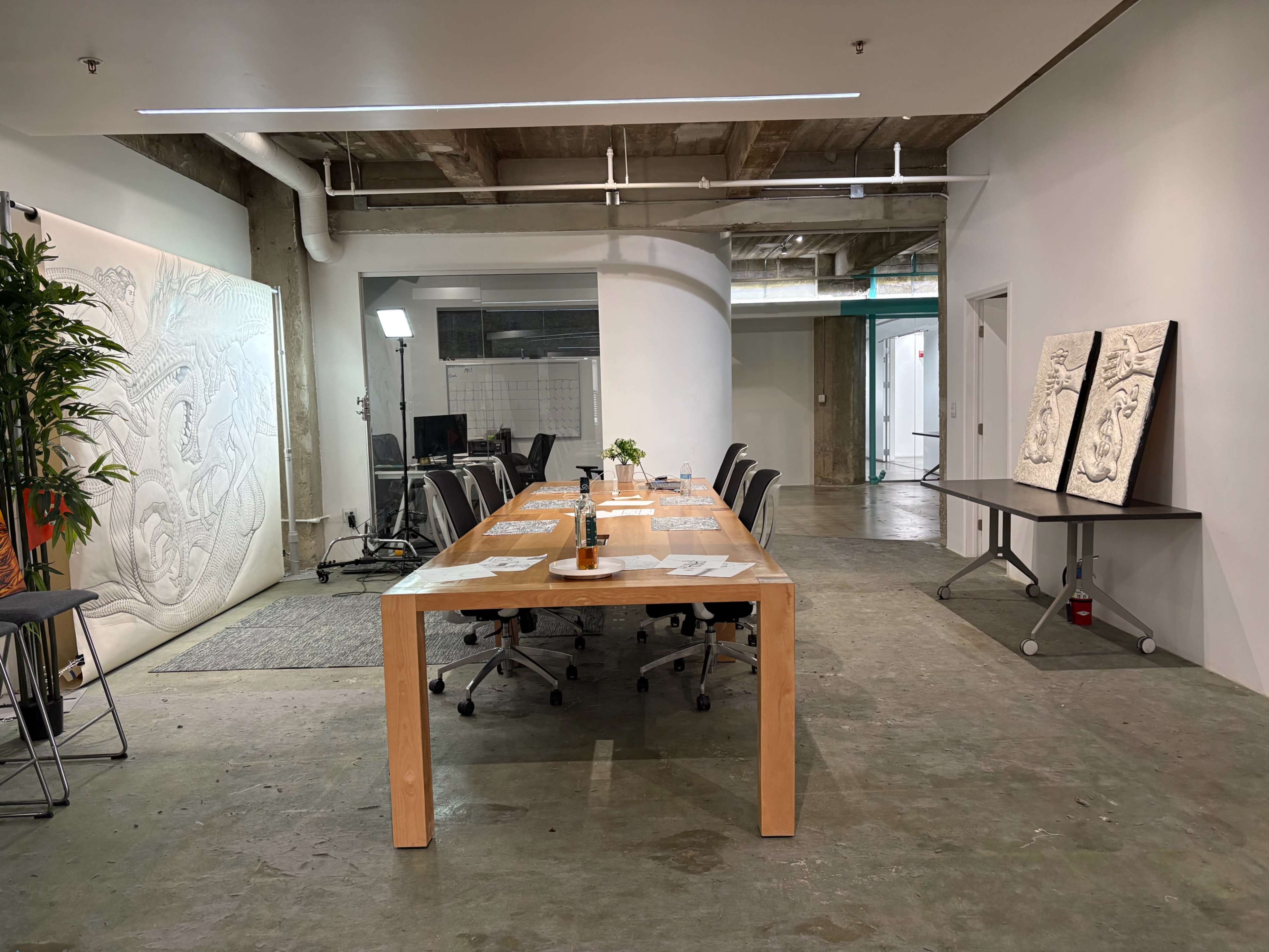 The image shows a modern conference room with a large wooden table surrounded by black chairs, artwork on the walls, and a small plant in the center.