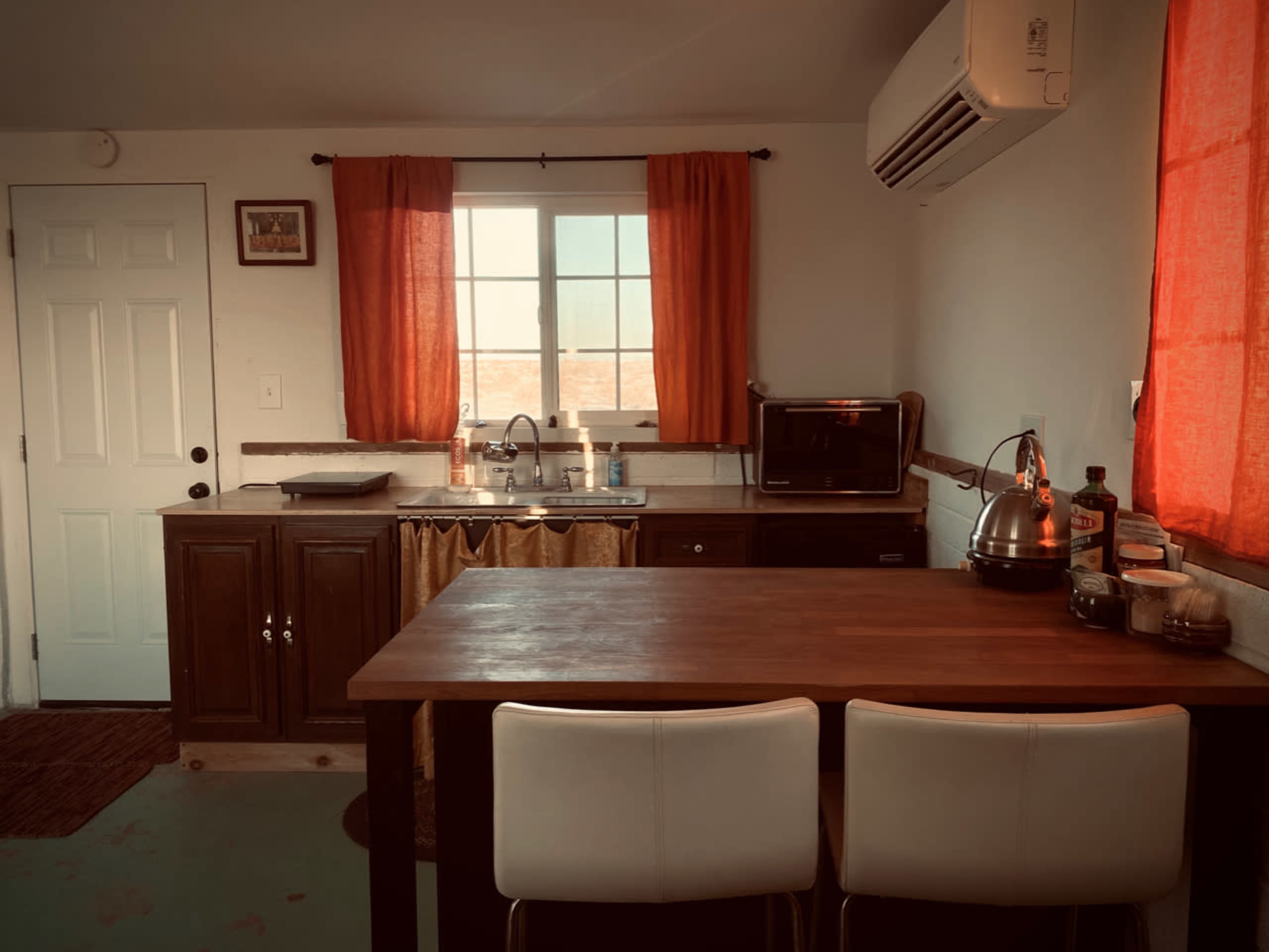 The image shows a small kitchen with a sink, microwave, and a wooden table with two white chairs, complemented by orange curtains at the window.