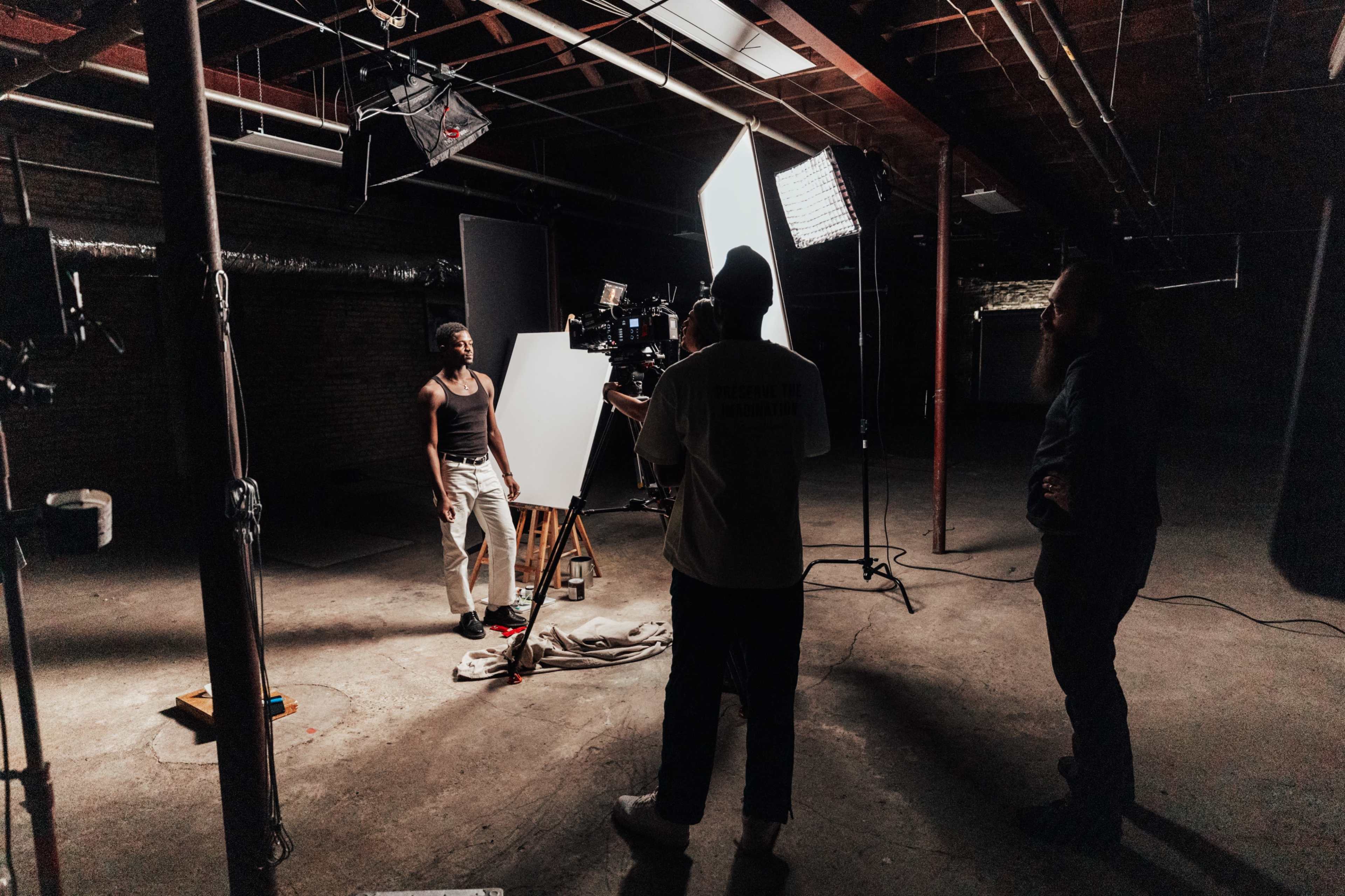A small film crew prepares to shoot a scene in a dimly lit industrial space with various lighting equipment set up.