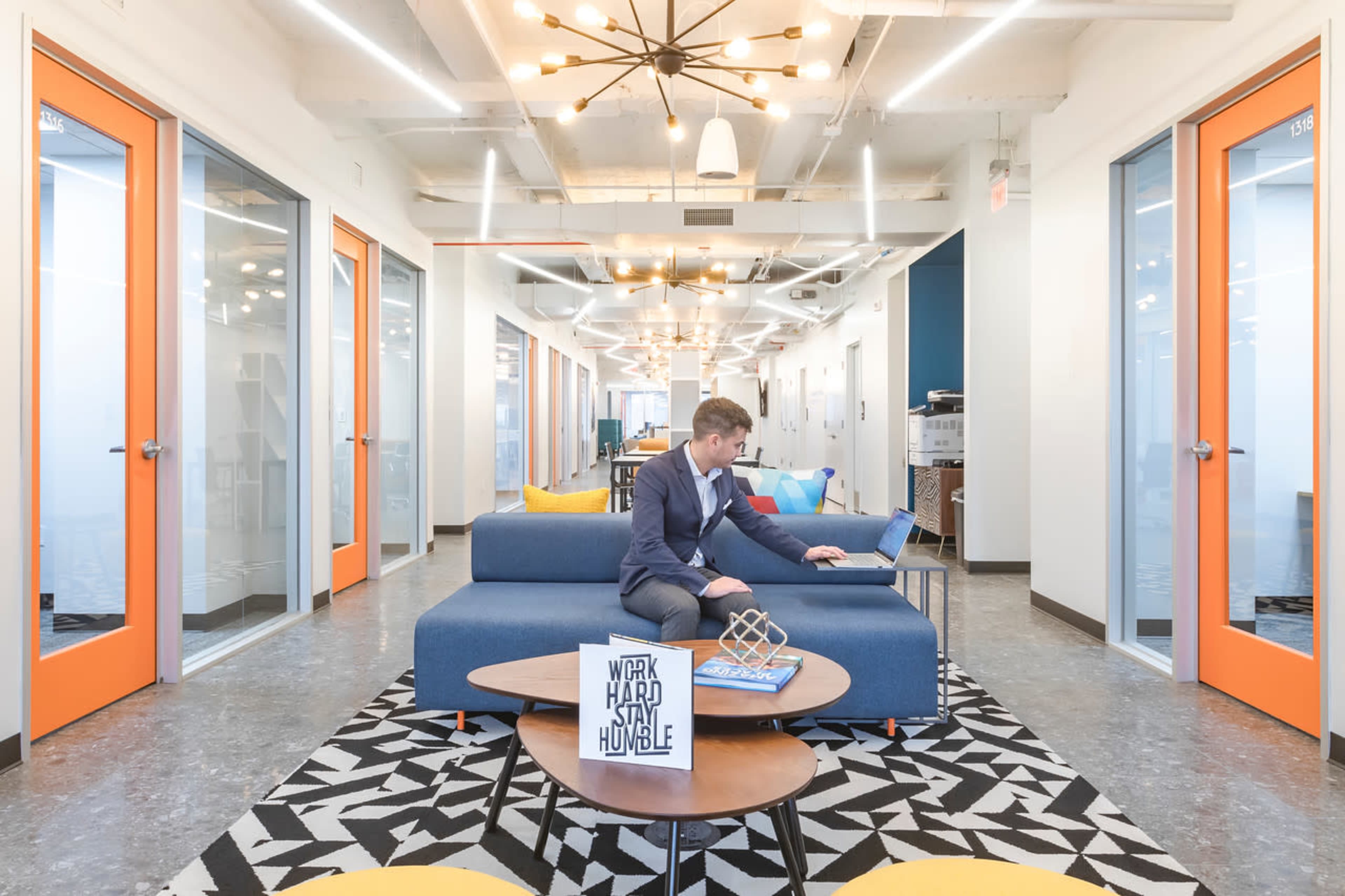 A man sits on a blue couch working on a laptop in a modern office space with bright orange doors and stylish lighting.