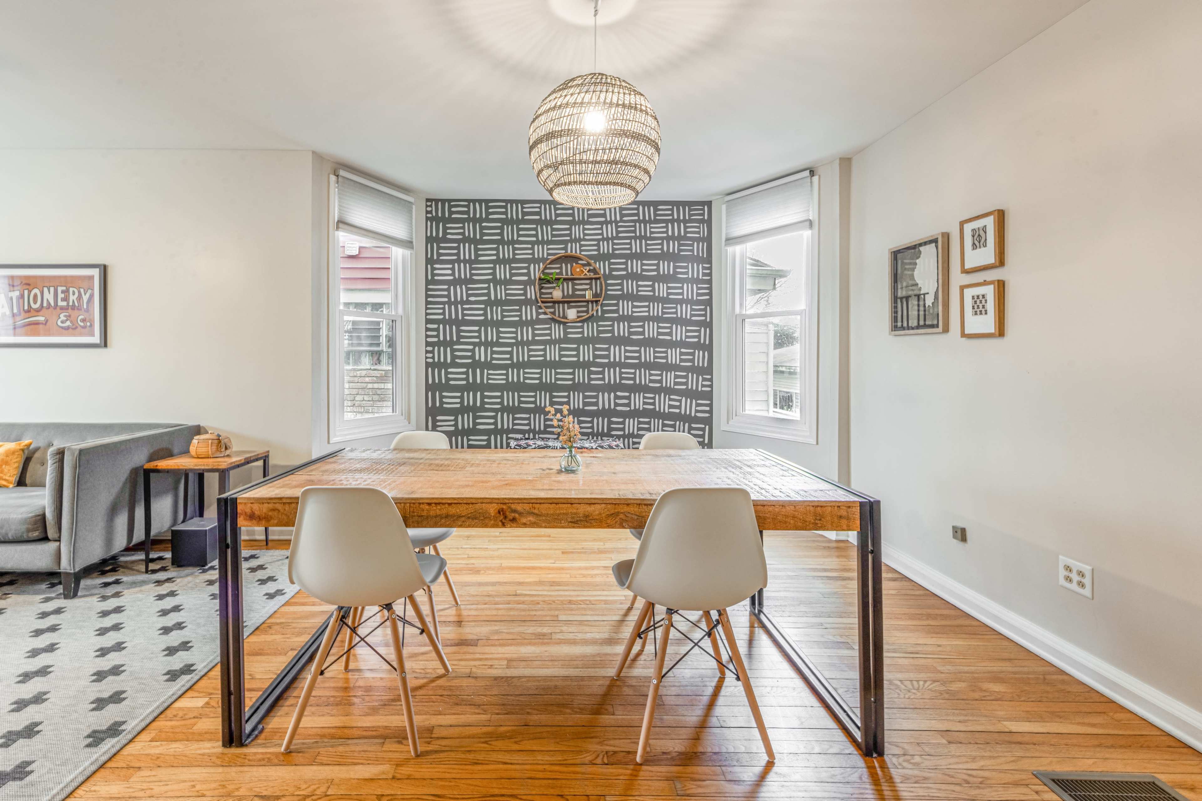 A modern dining area features a wooden table with metal legs and four white chairs, surrounded by large windows and a decorative wall pattern.