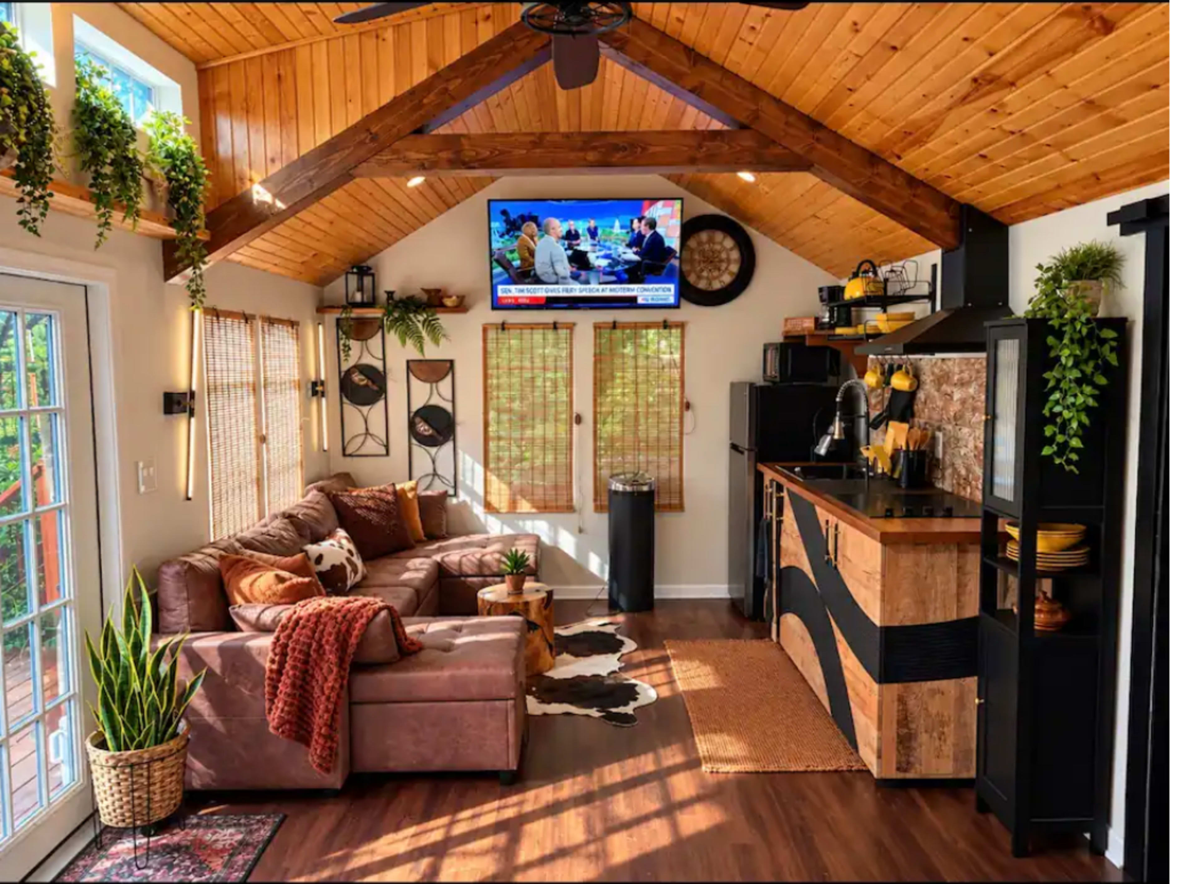 The image shows a cozy interior of a small living space with a brown sofa, a kitchen area, and a television mounted on the wall, illuminated by natural light from large windows.