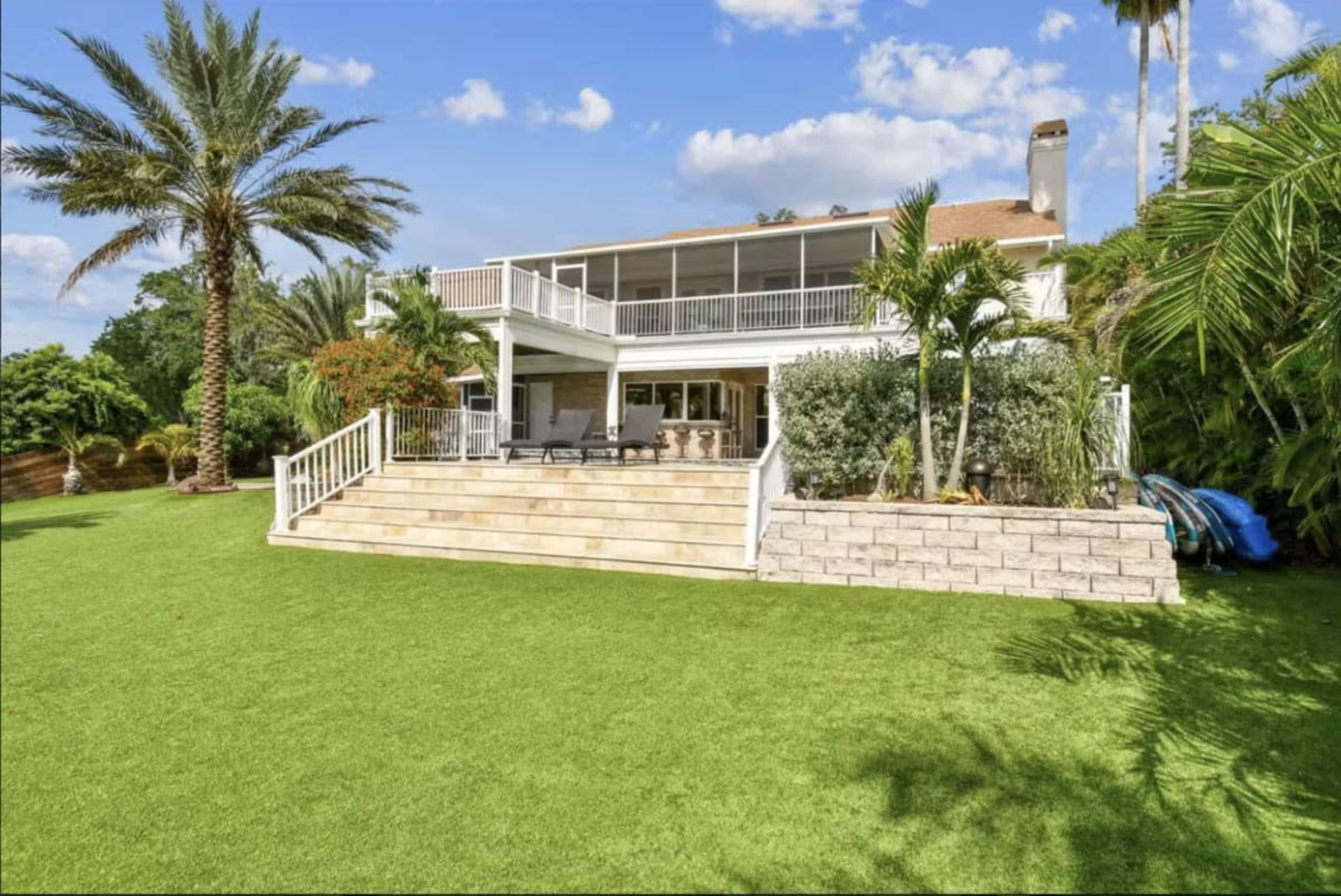 A two-story house with a large backyard features a green lawn, palm trees, and a patio area with outdoor seating.