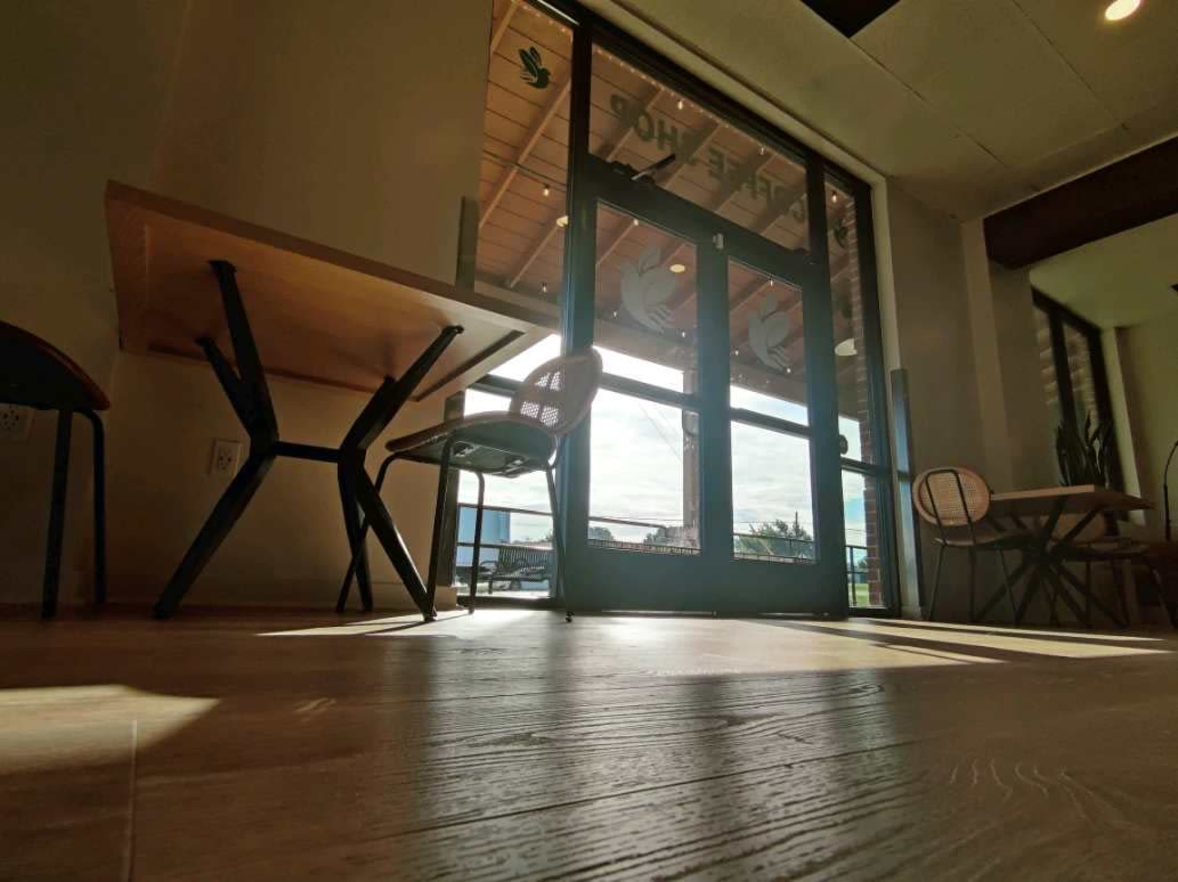 The image shows the interior of a café with large glass doors leading to the outside, a wooden table, and chairs arranged nearby.