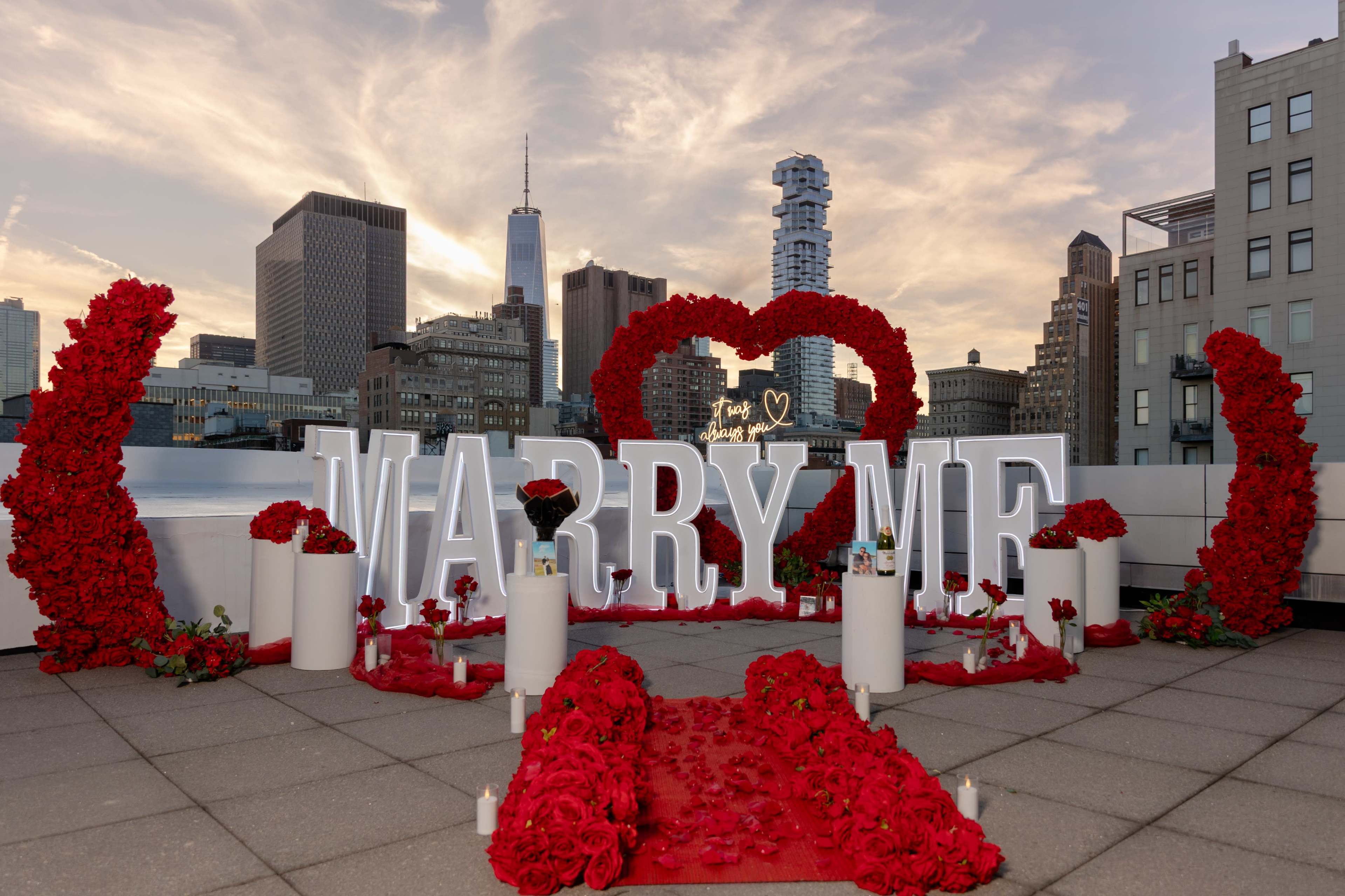 A romantic rooftop proposal setup features large "MARRY ME" letters surrounded by red roses, candles, and a heart-shaped arrangement, with a city skyline in the background.