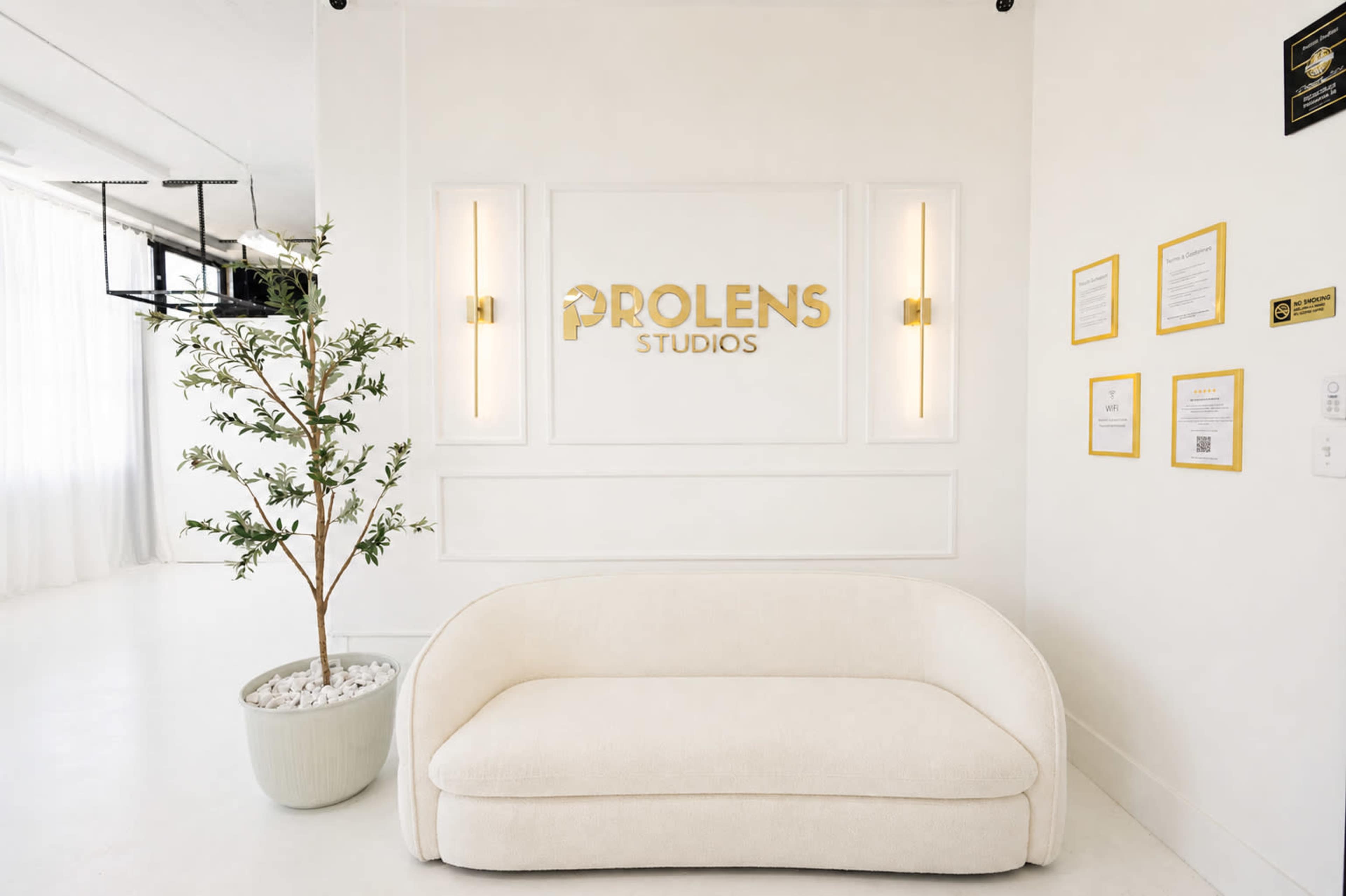 The image shows a minimalist studio reception area with a white couch, a potted plant, and a prominently displayed sign that reads "PROLENS STUDIOS."