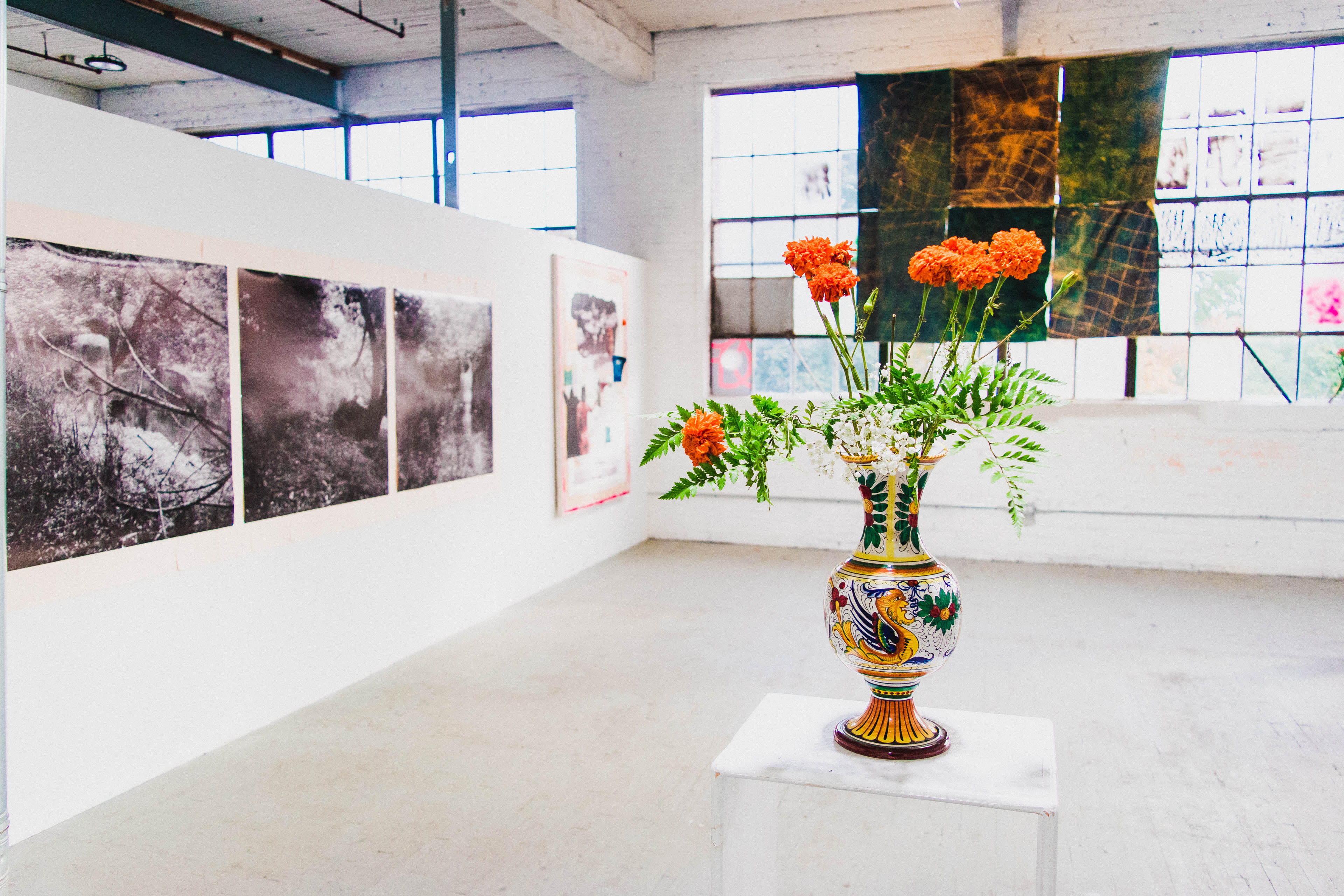 The image shows an art gallery with framed black-and-white photographs on the walls and a colorful vase of flowers on a pedestal in the foreground.