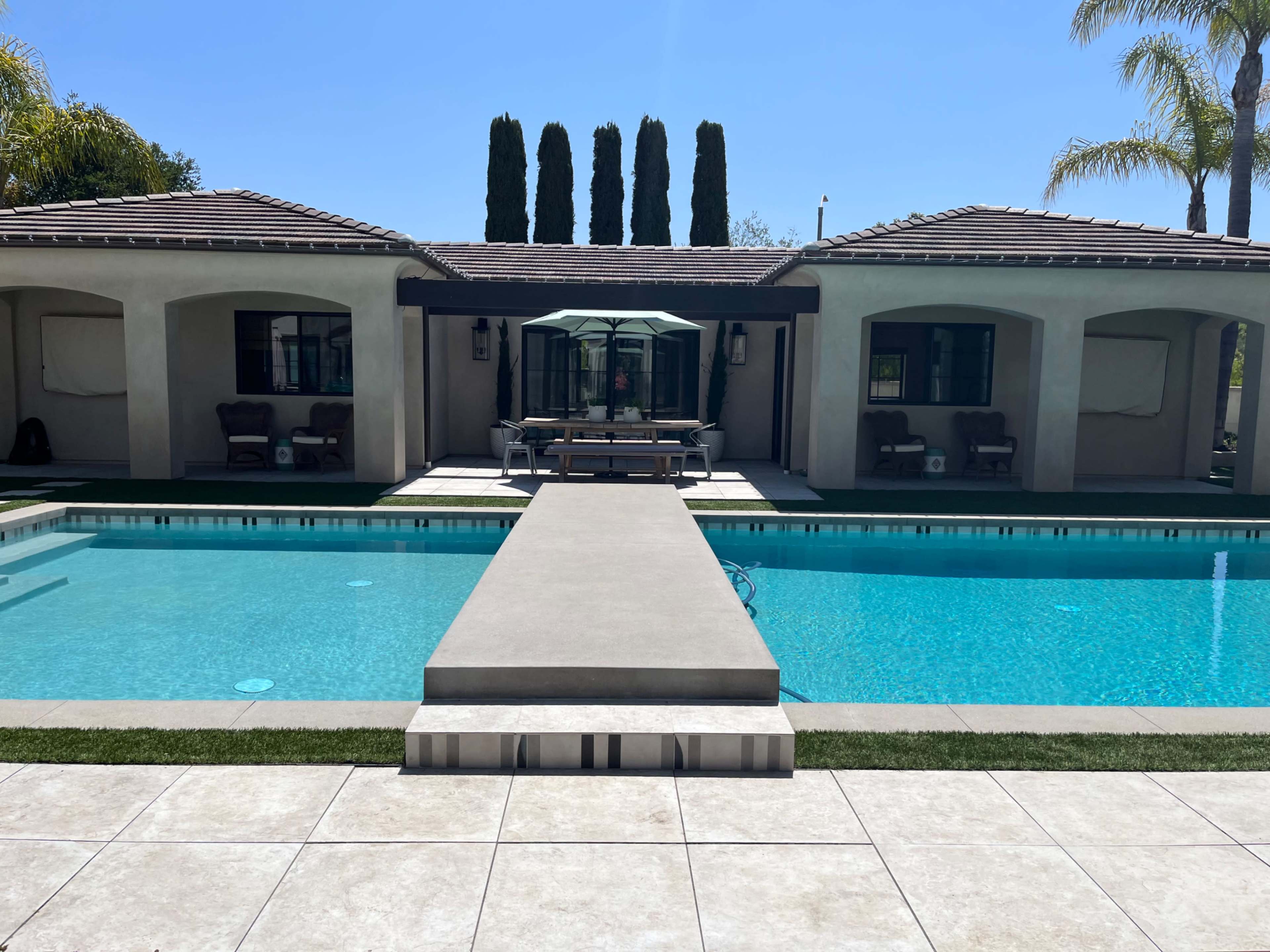 A spacious swimming pool with a walkway leading to a house featuring two covered patios and palm trees in the background.