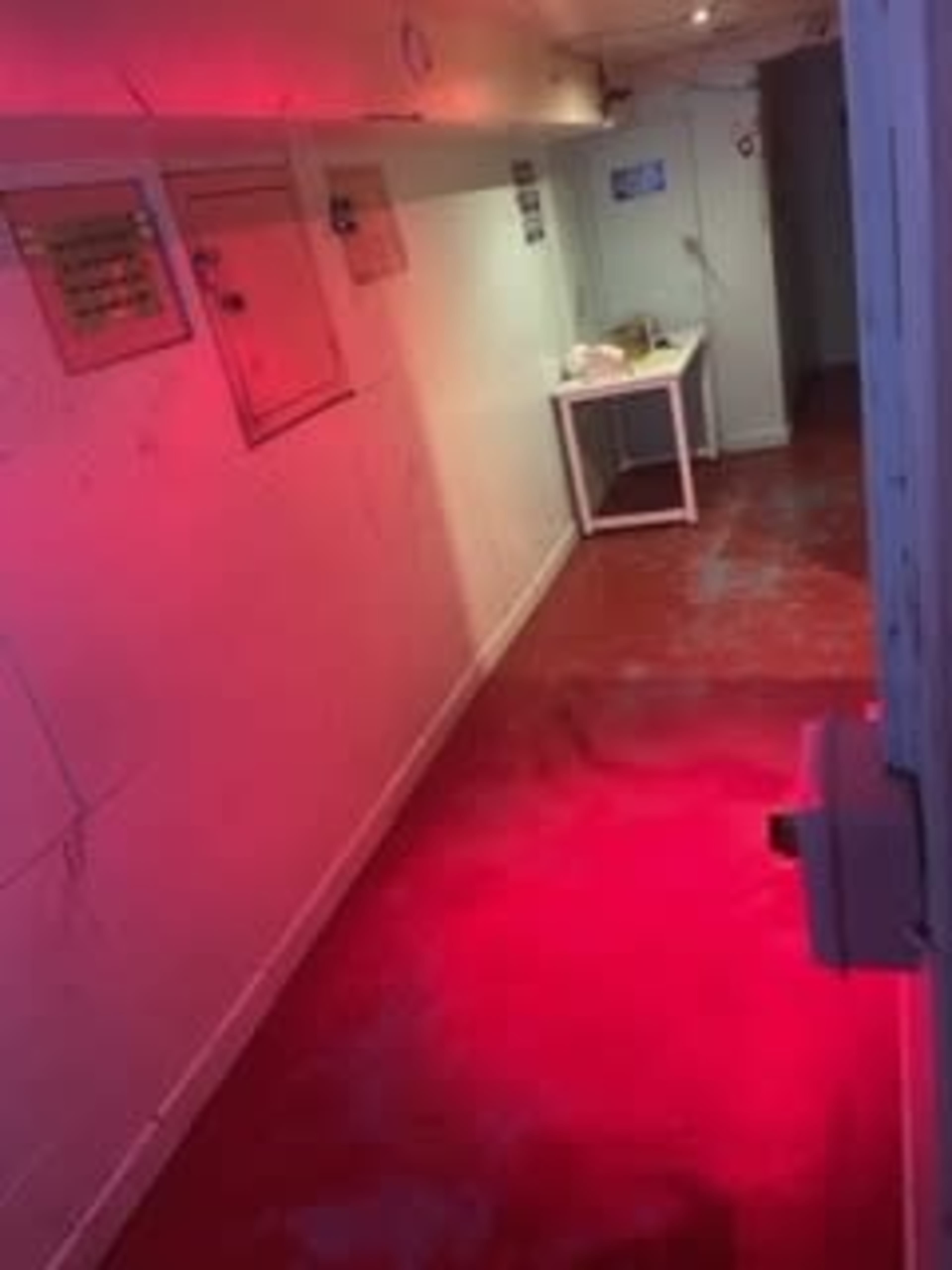 A narrow hallway with red lighting, white walls, and a table at the end.