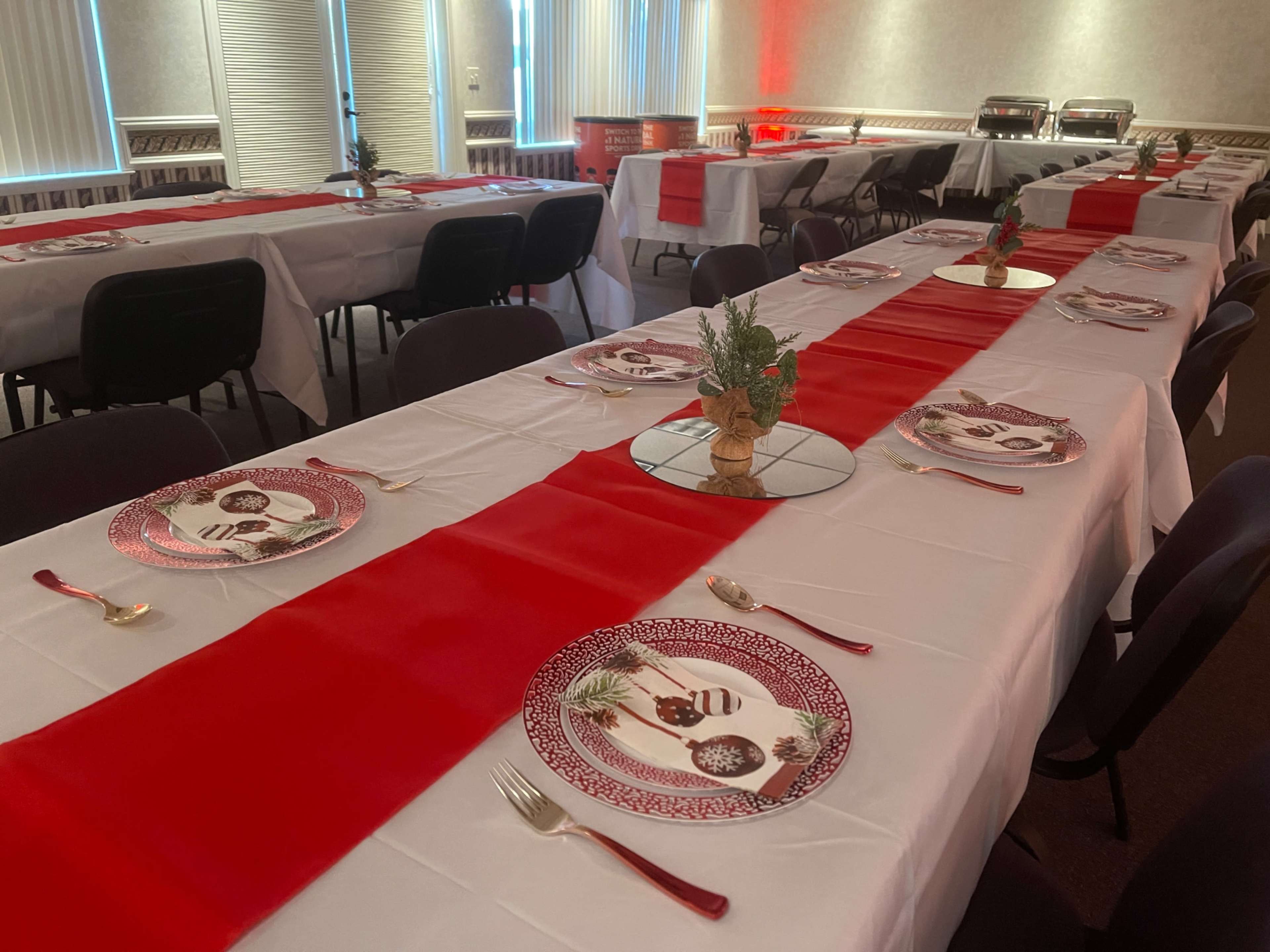 A dining setup features multiple tables covered with white tablecloths and red runners, arranged with decorative plates and utensils.