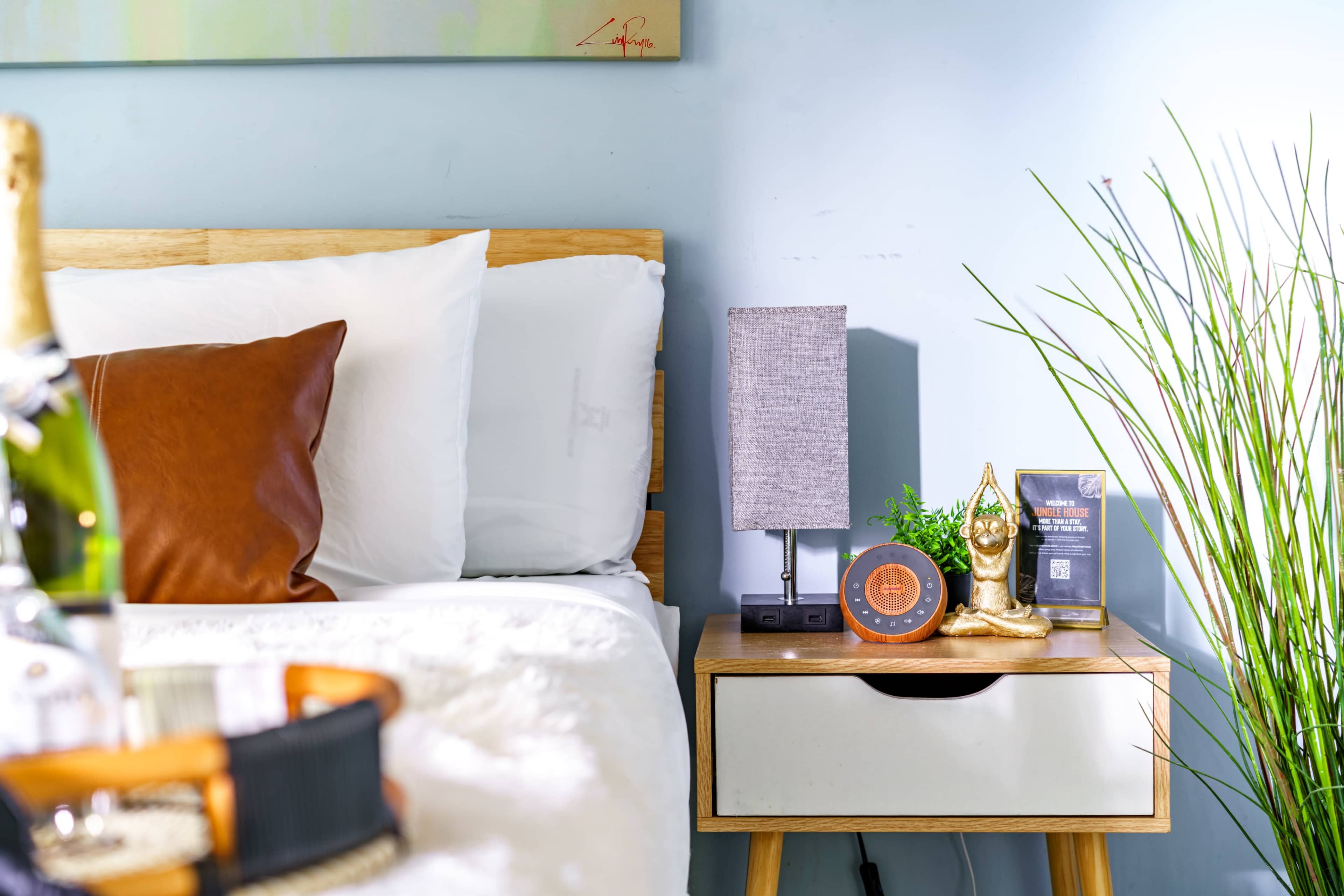 A neatly arranged bedroom features a wooden bedside table with a lamp, a decorative statue, and a bottle of champagne on the bed.