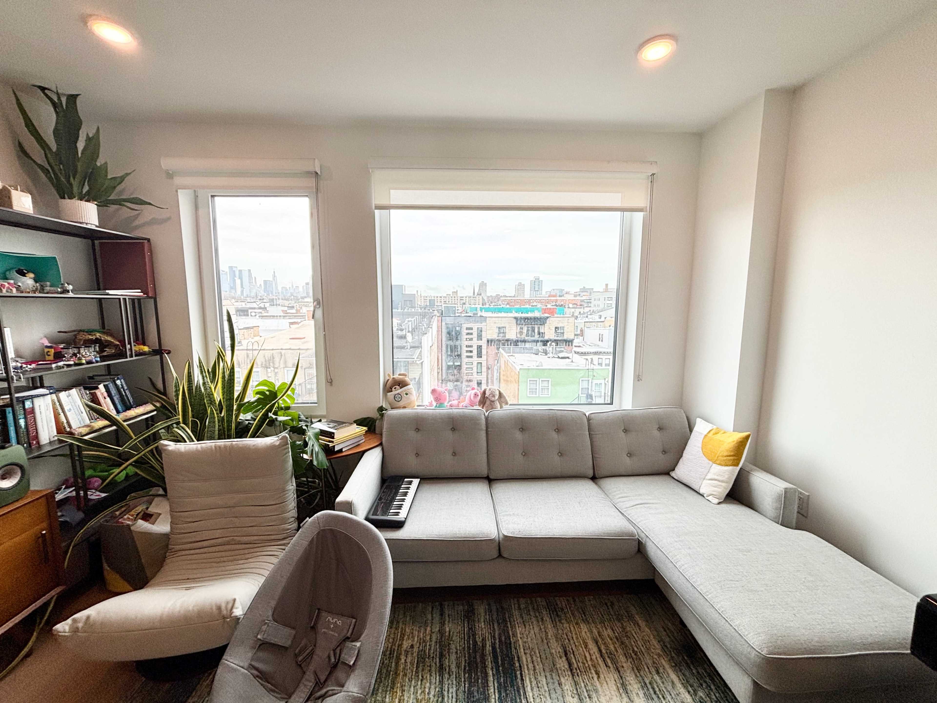 The image shows a modern living room with a sectional sofa, a music keyboard, a plant in the corner, and large windows offering a view of a cityscape.