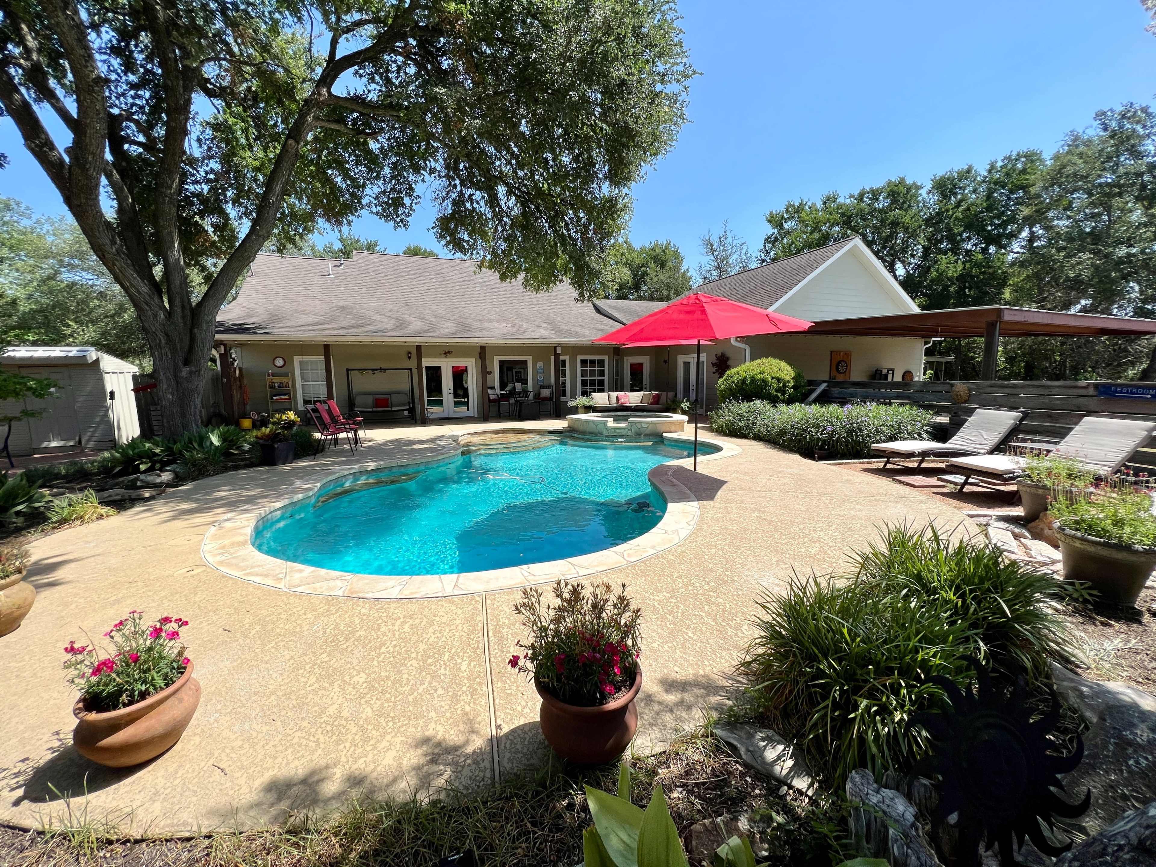 A backyard features a swimming pool surrounded by lounge chairs, potted plants, and an umbrella, with a house and trees in the background.