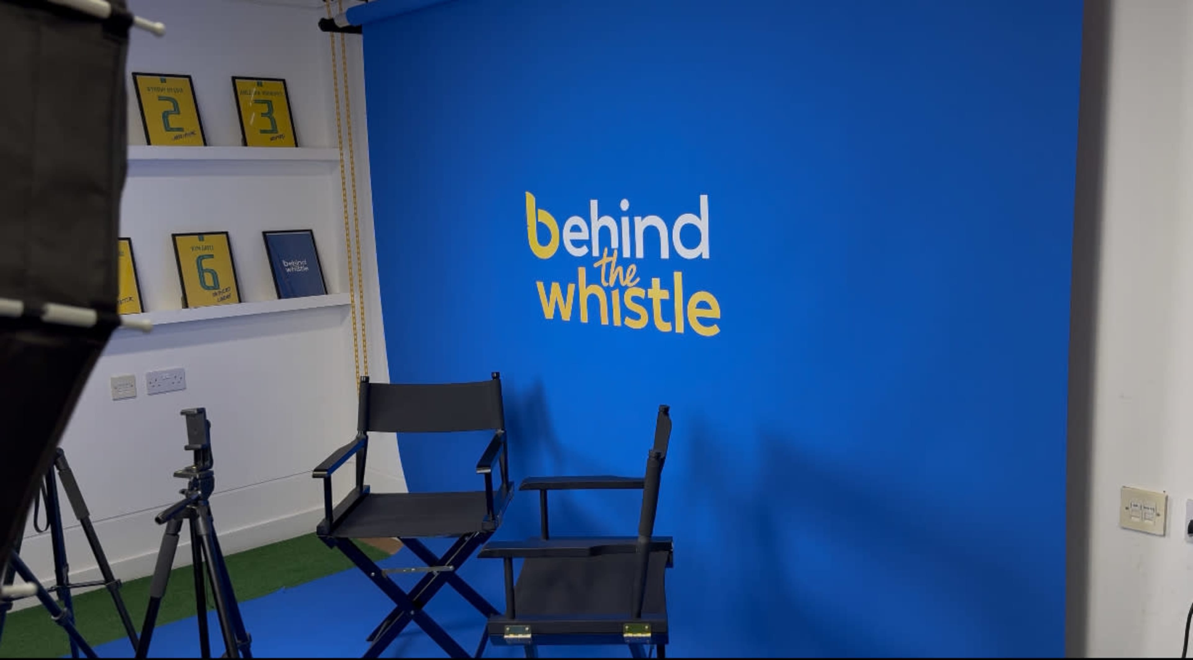 The image shows a studio setup with a bright blue backdrop featuring the text "behind the whistle," along with two empty director's chairs and some framed items on a shelf.