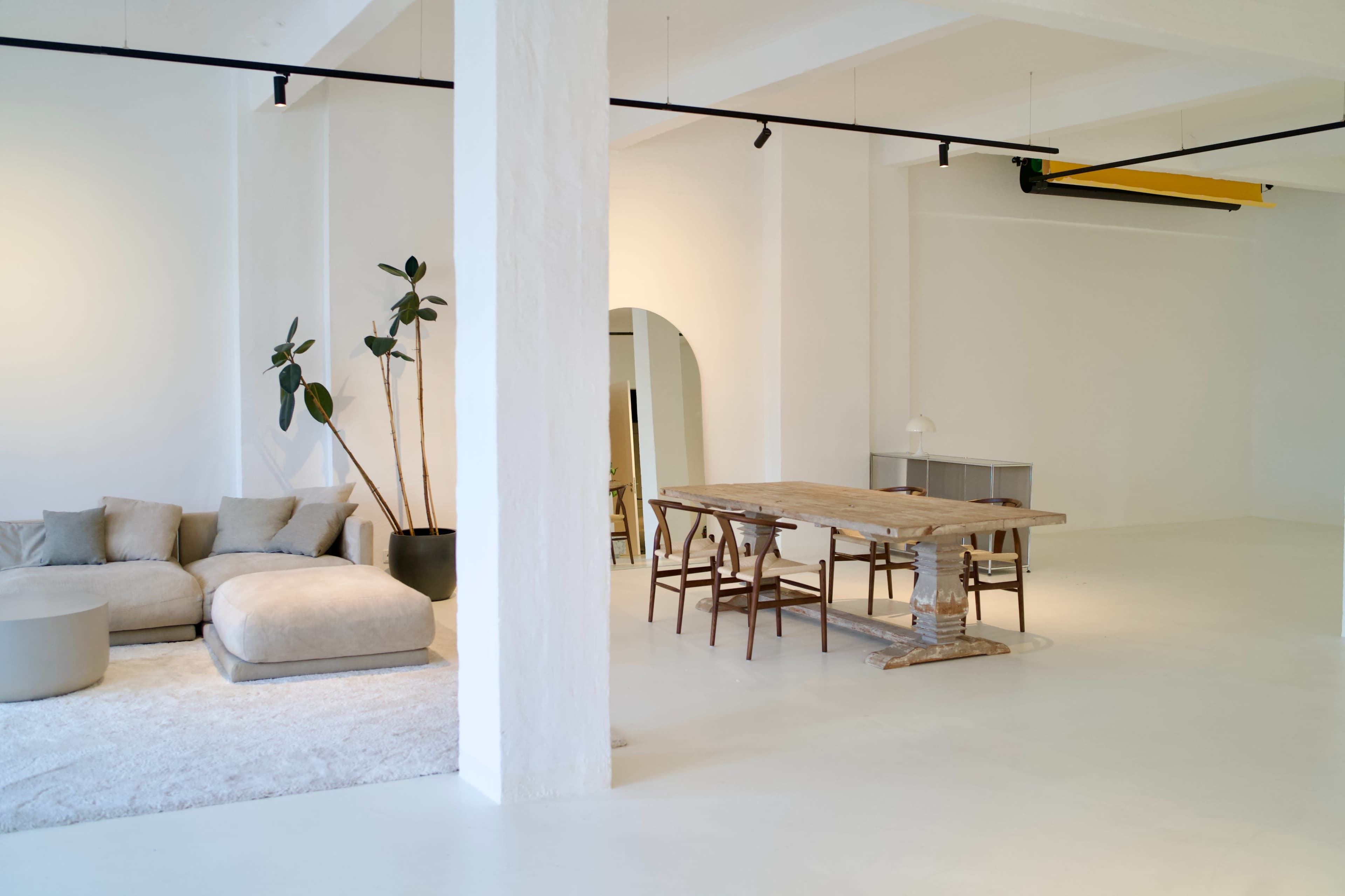 The image shows a spacious, minimalistic interior featuring a large sofa, a wooden dining table, and potted plants against white walls.