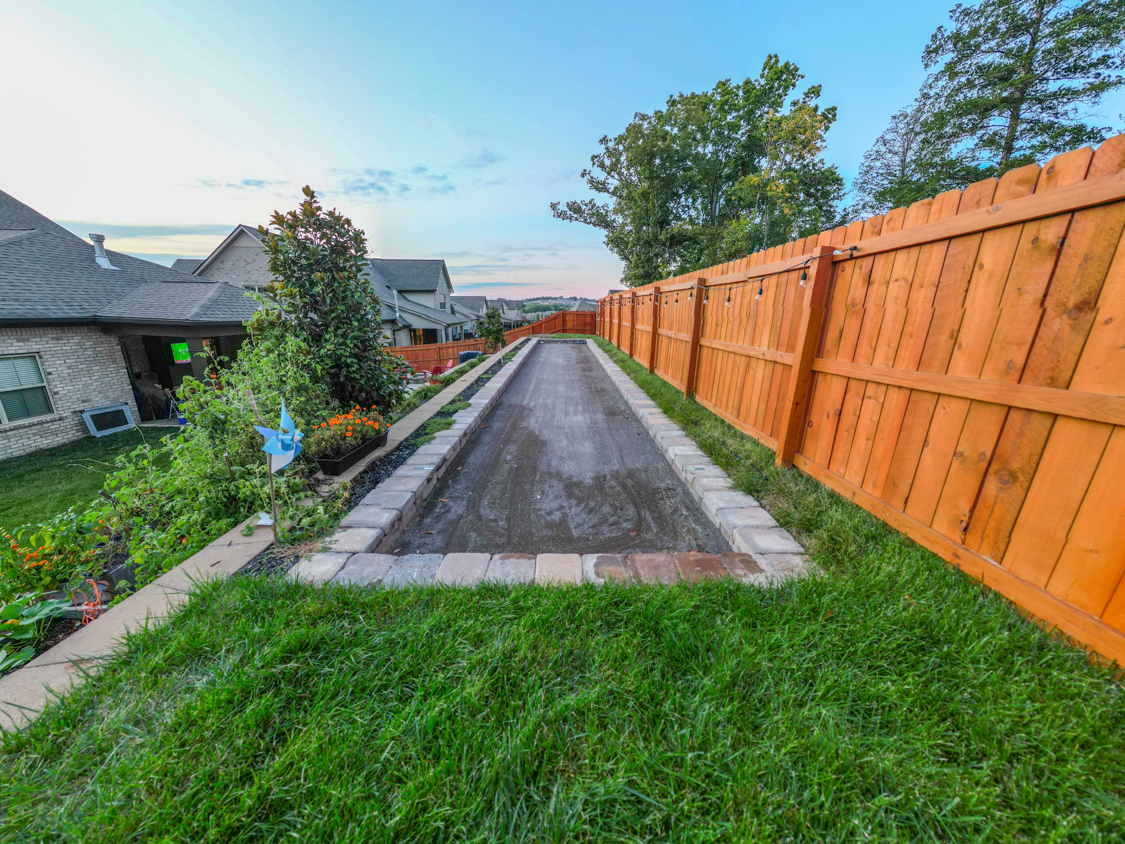 The image shows a narrow gravel path bordered by a stone edge, flanked by a tall wooden fence and grassy areas on either side.